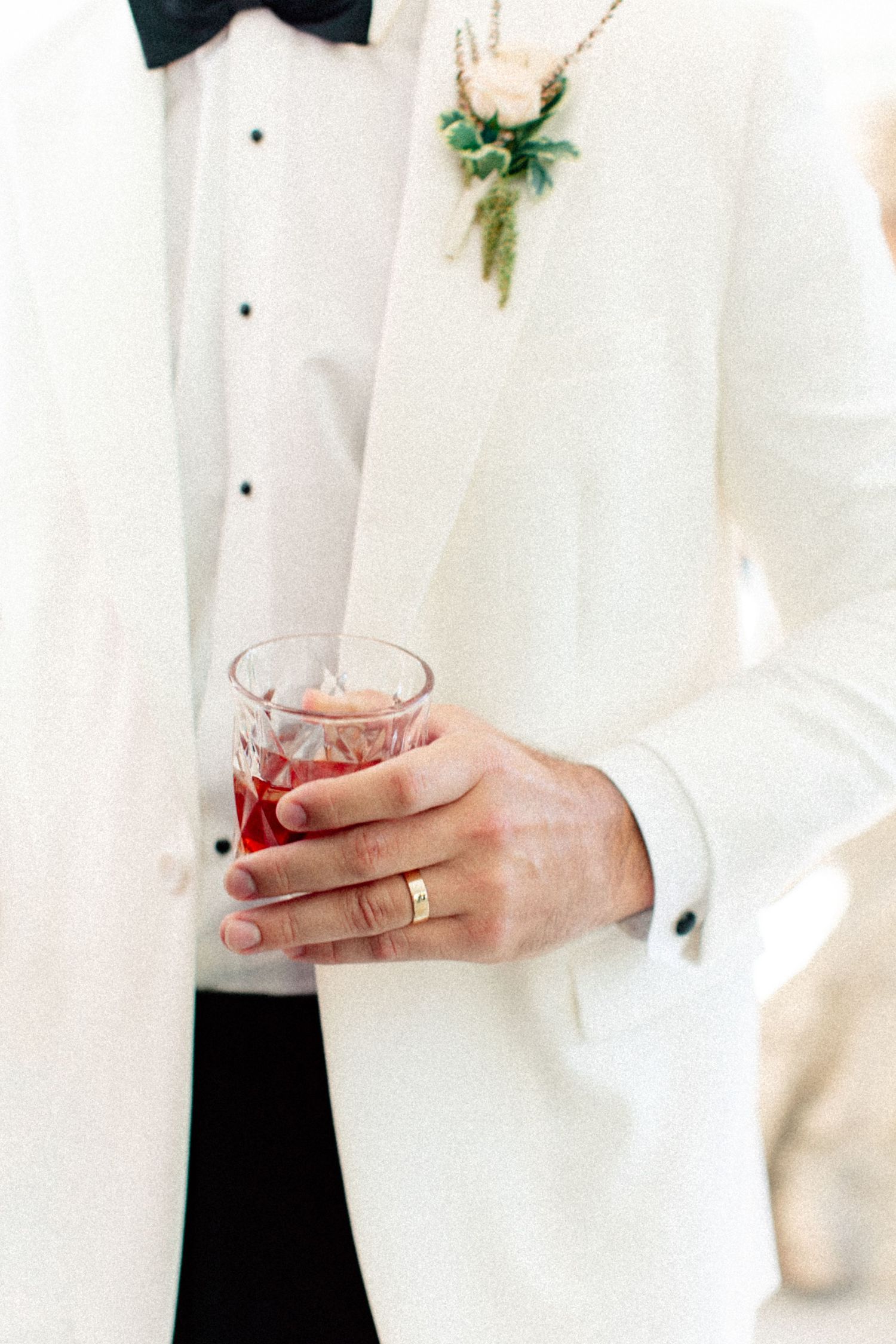 Groom in white tuxedo jacket with floral boutonniere holding a crystal glass of red drink, wearing a gold wedding band.