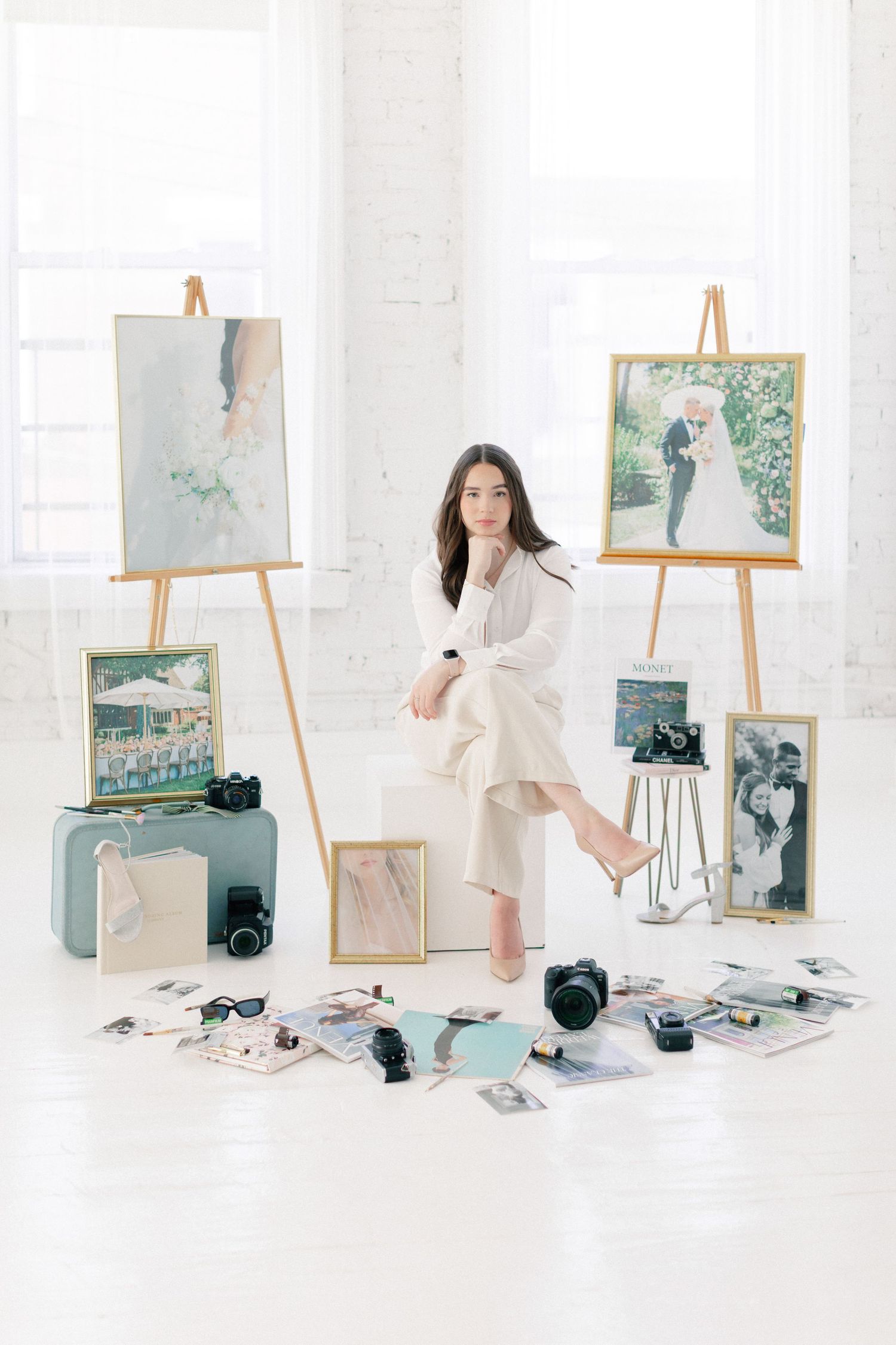 Young female artist sits surrounded by easels with paintings, cameras, and scattered photos in a bright white studio space.