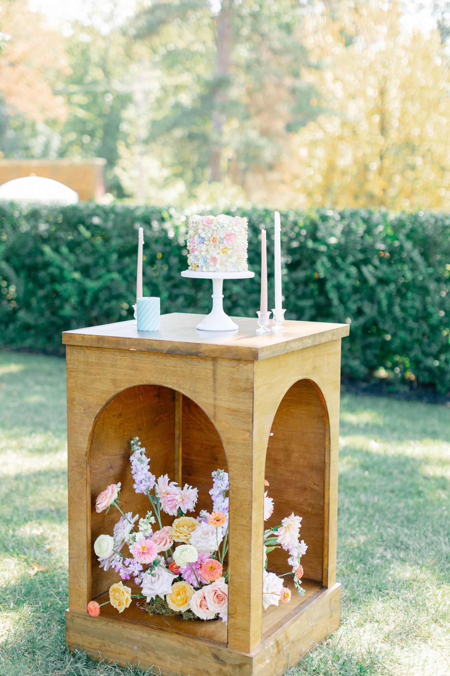 Rustic wooden display stand with small white wedding cake, taper candles, and colorful floral arrangement underneath.