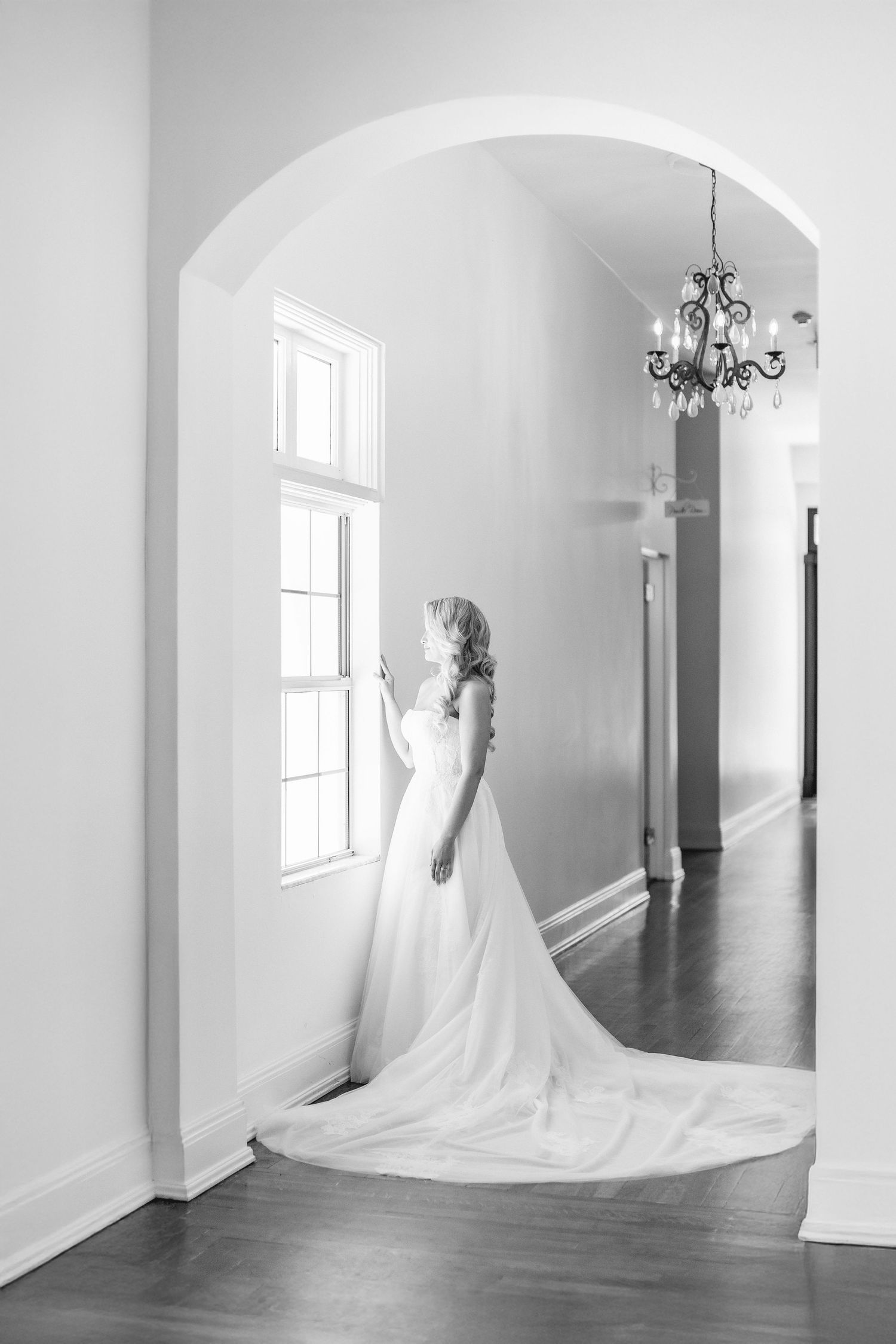 Bride in a flowing white gown with train stands by a window under an arch, with a chandelier in a black and white photo wedding at Bella Cos