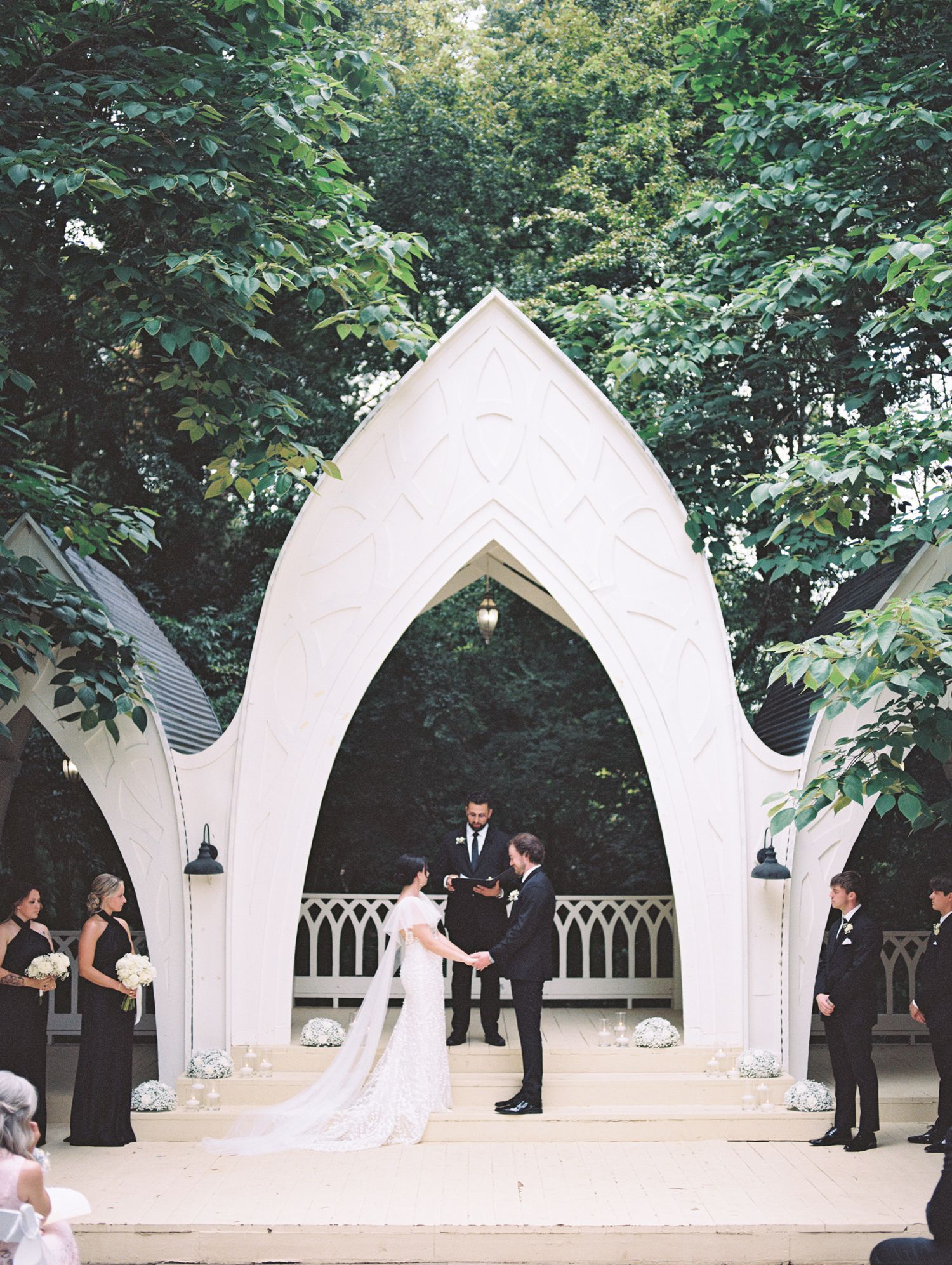 Outdoor wedding ceremony under a white Gothic arch with bride and groom holding hands, flanked by wedding party amid lush trees.