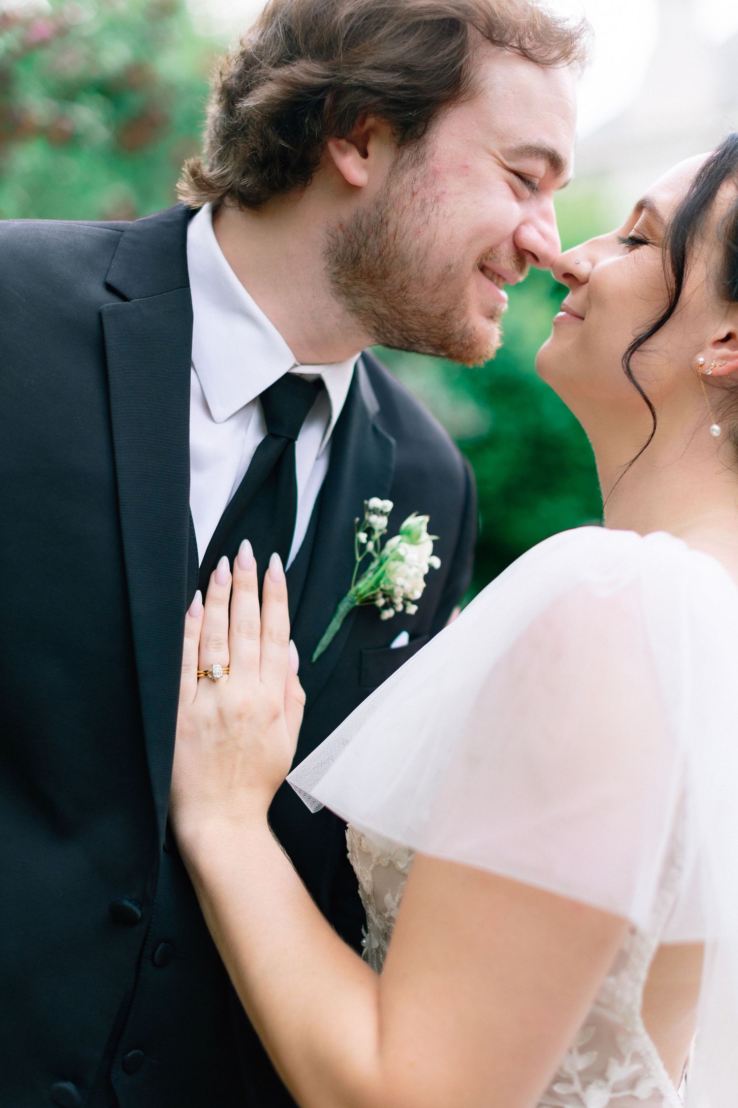 A smiling bride and groom share an intimate moment, nearly touching noses, as she displays her wedding ring on his chest.