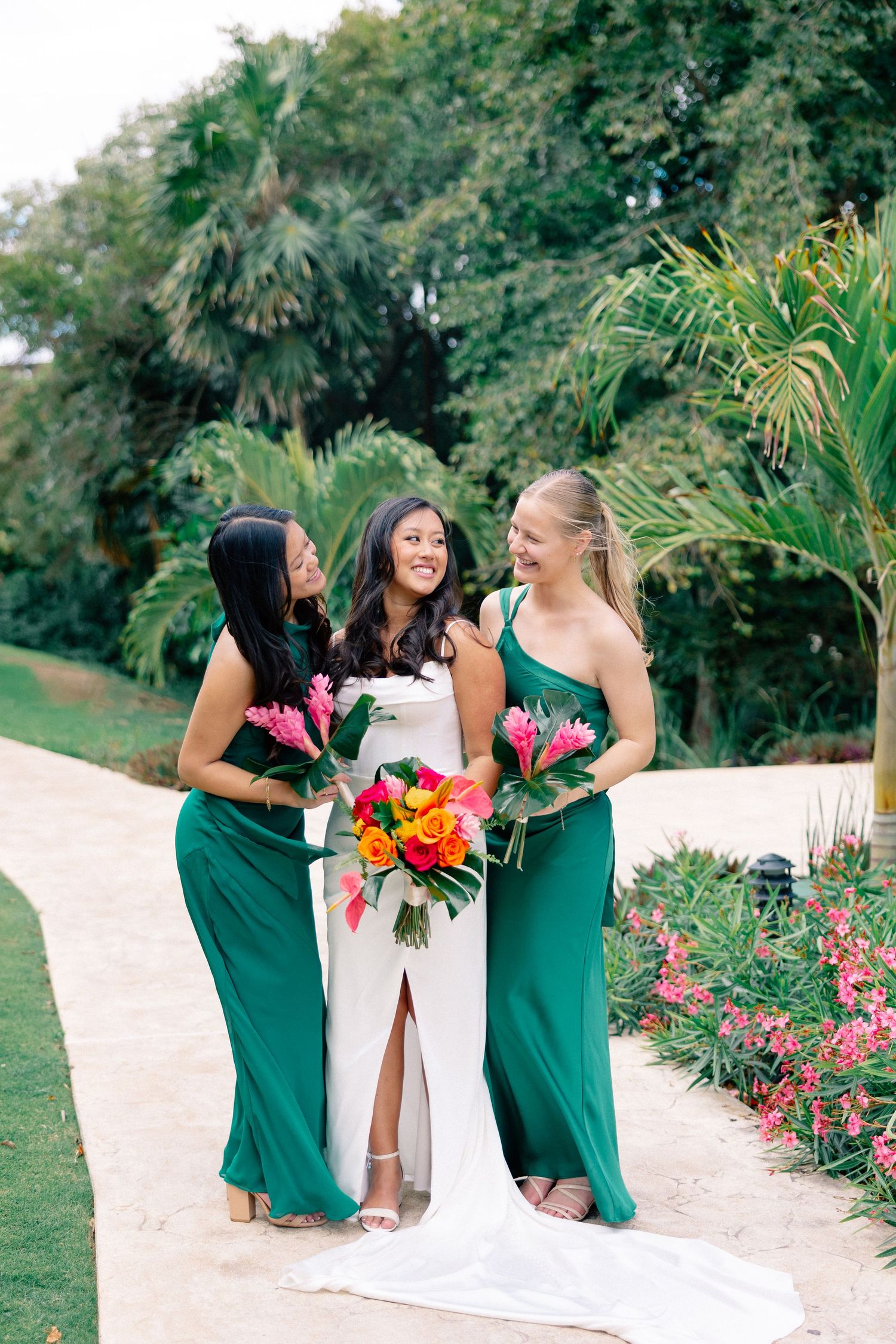 Bride in white gown with two bridesmaids in emerald green dresses holding tropical bouquets outdoors.