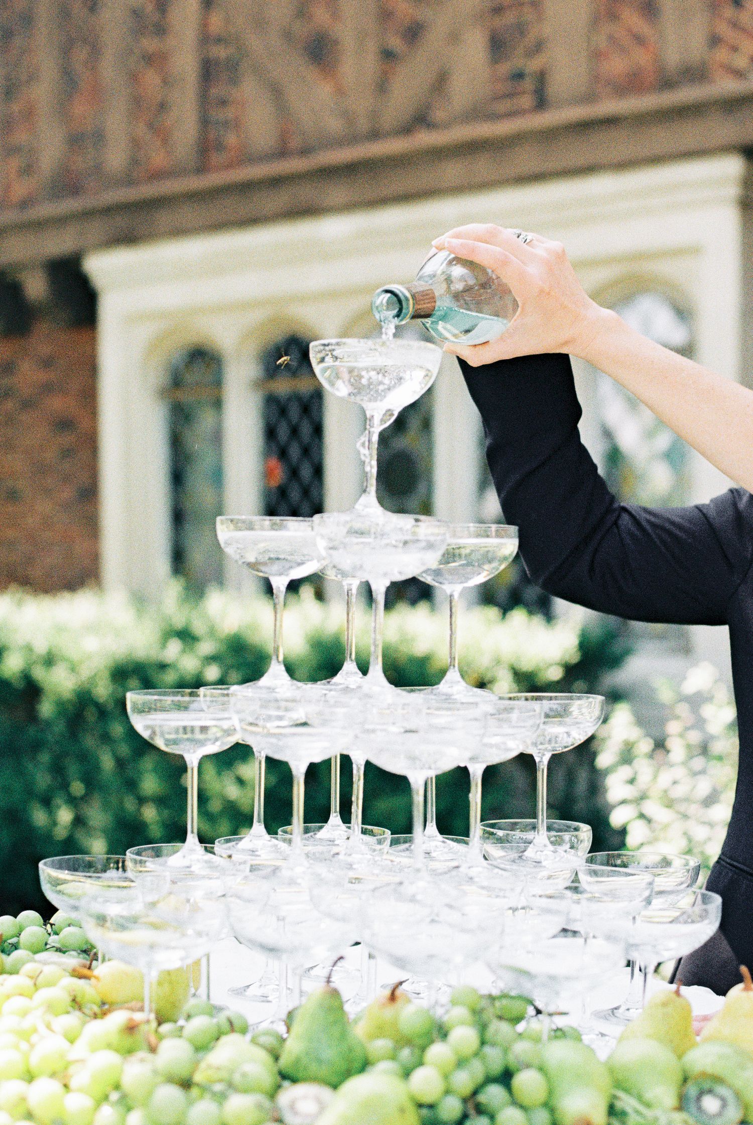 Champagne tower being filled at outdoor wedding reception surrounded by fresh green grapes and pears.