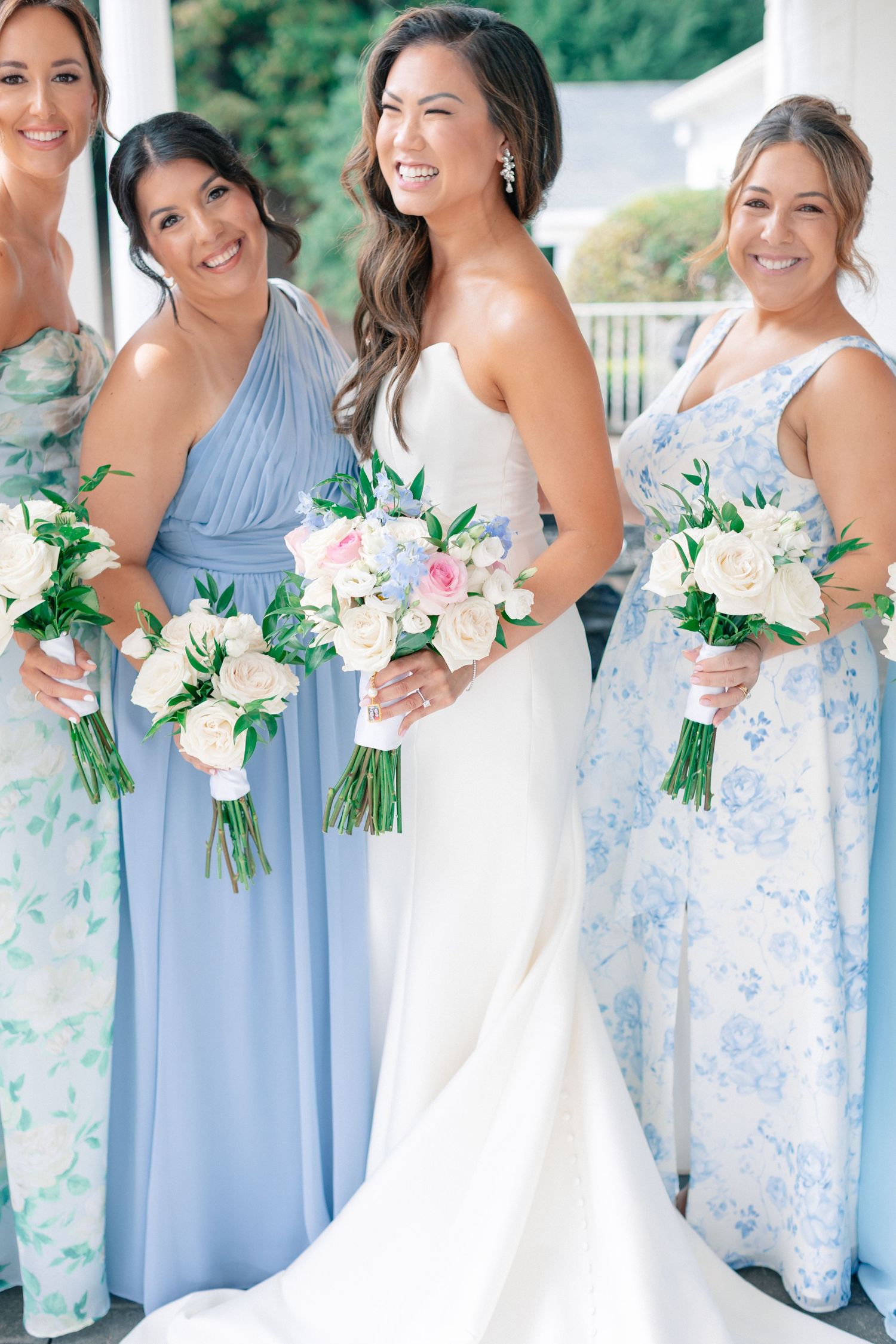 Bride in white gown poses with bridesmaids in blue floral dresses holding white and pink bouquets.
