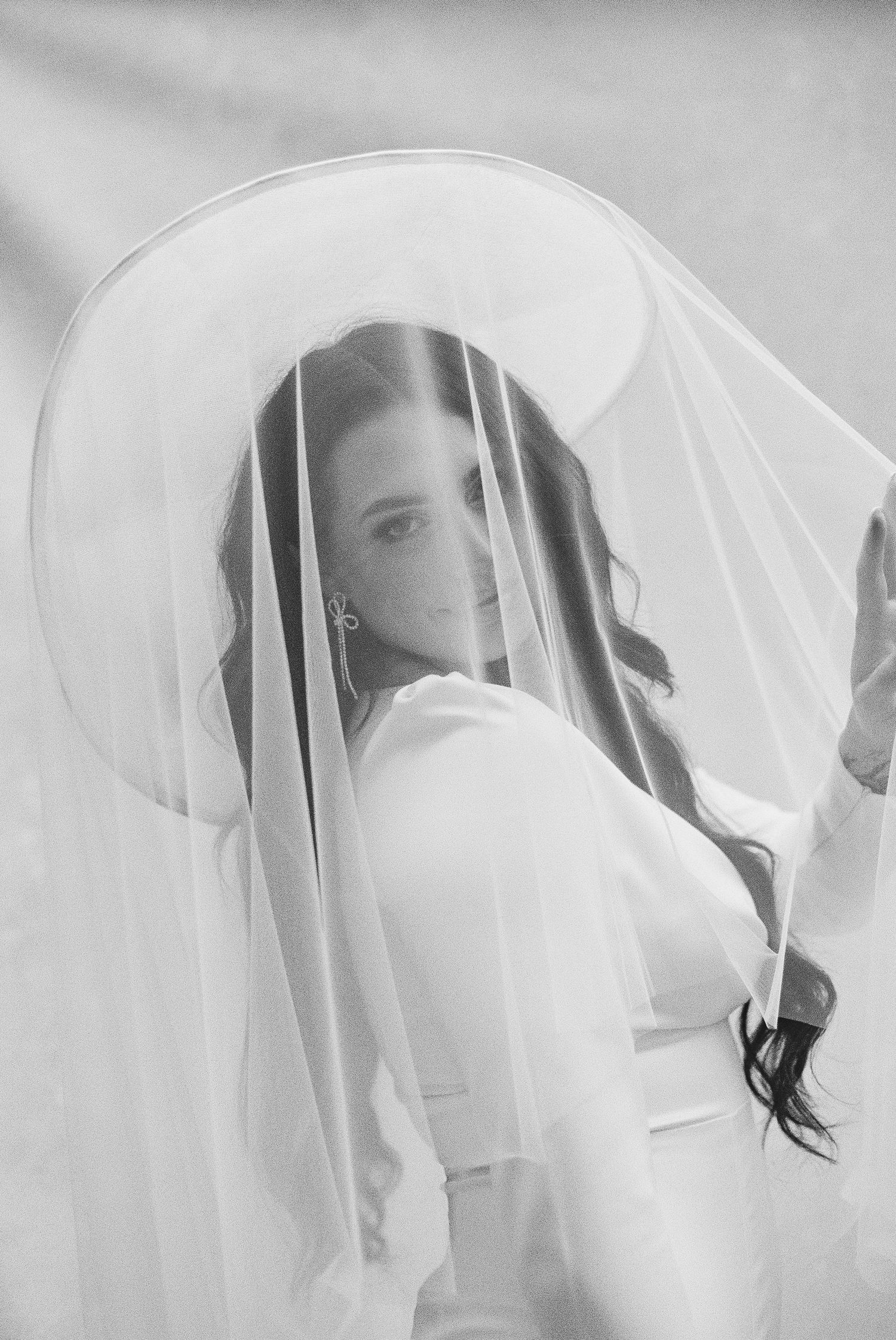 Black and white bridal portrait of a woman wearing a wide-brim hat with a sheer veil draped over her face.