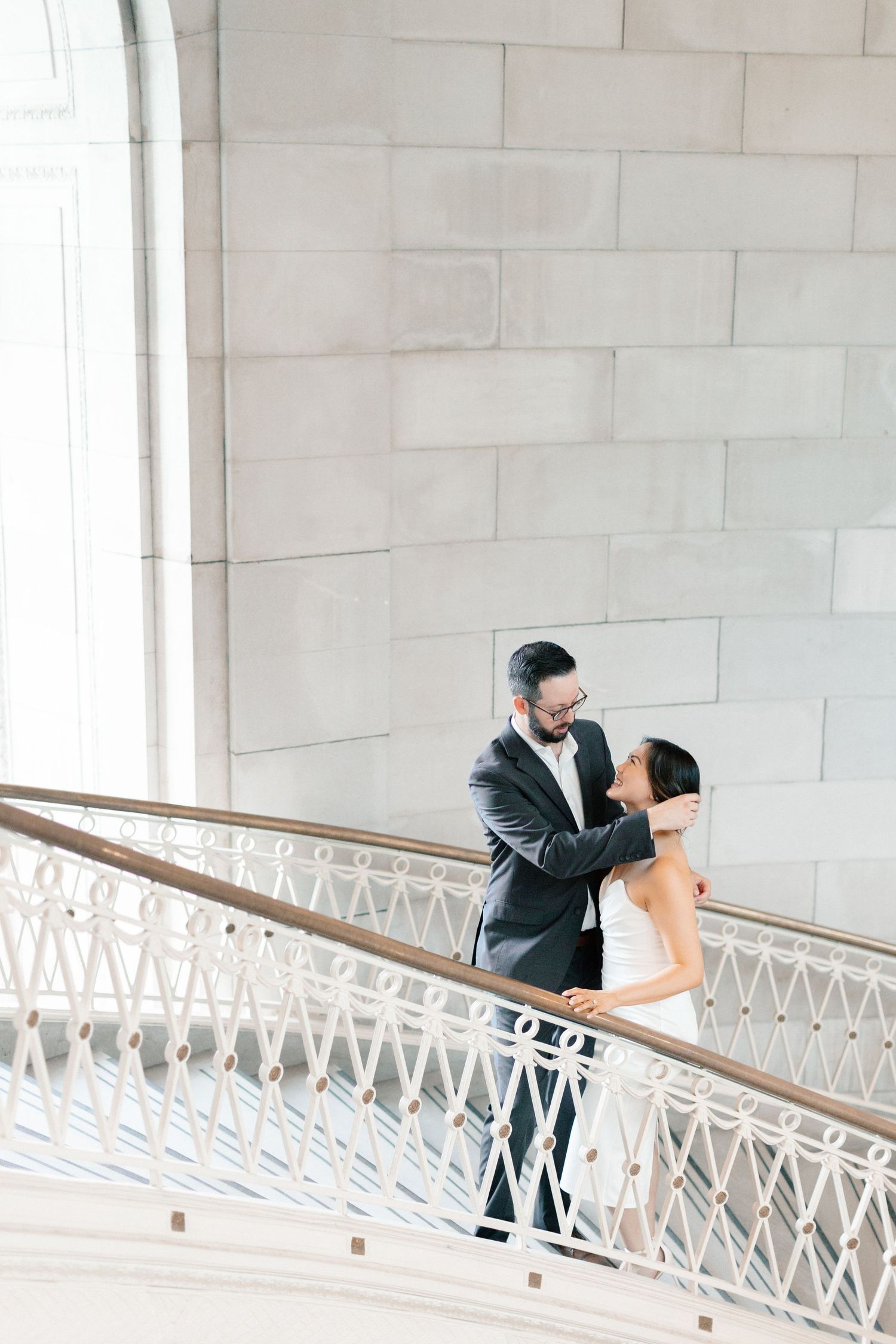 Bride and groom share a romantic embrace on an elegant white spiral staircase with ornate iron railings.