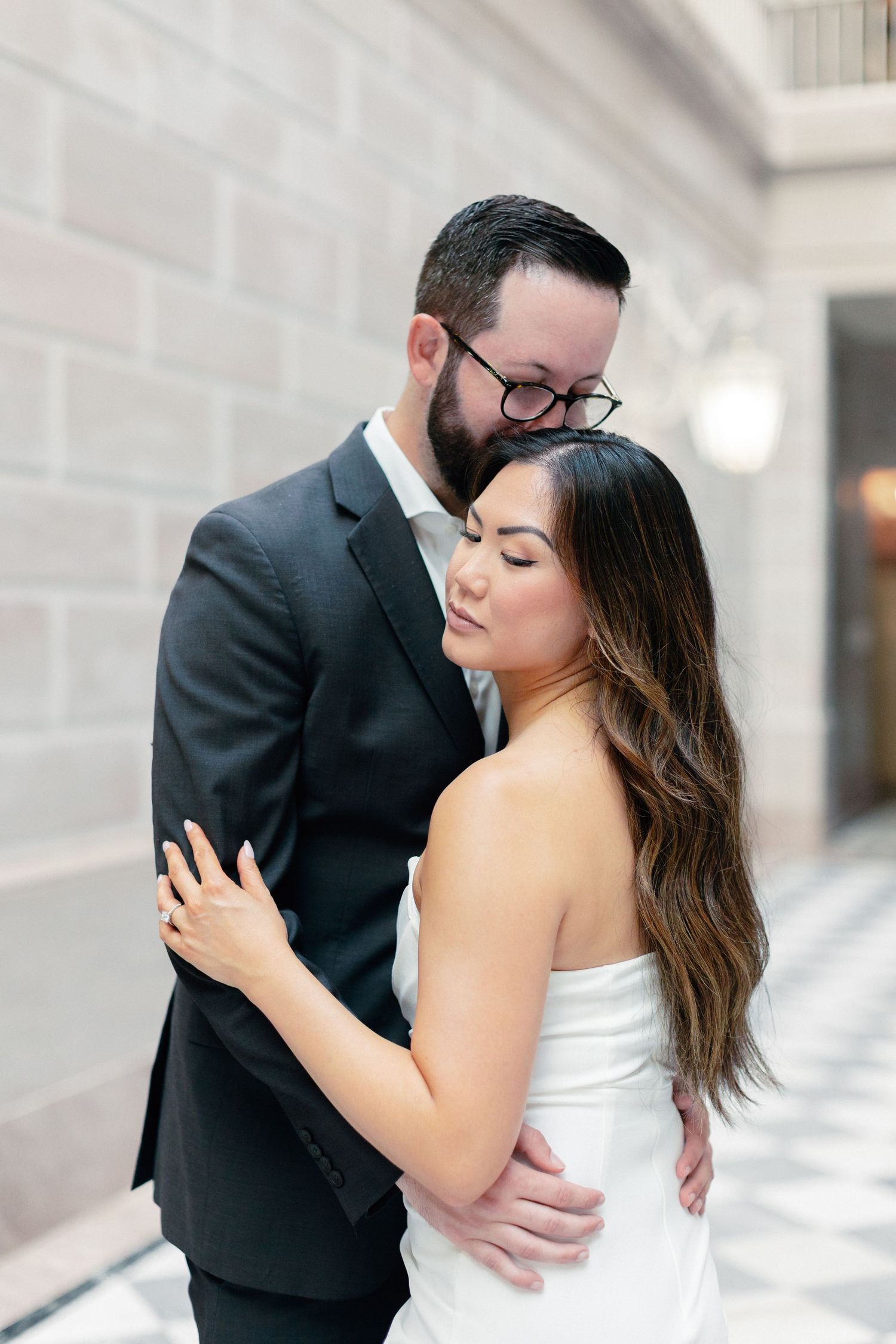 A groom in a dark suit embraces his bride in a white strapless gown, sharing a tender moment in an elegant hallway.