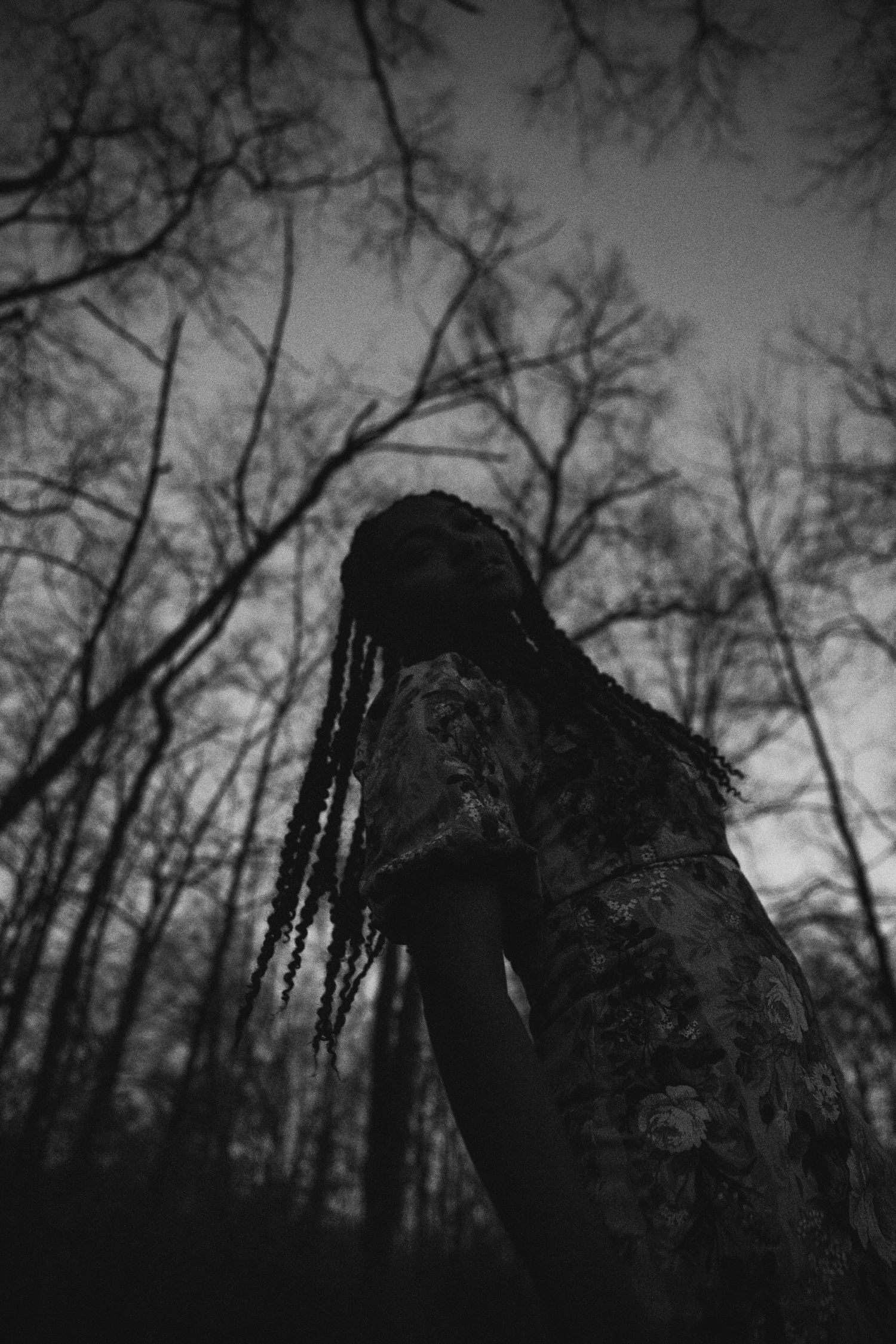 Black and white photo of person with dreadlocks in dark forest with bare trees at dusk.
