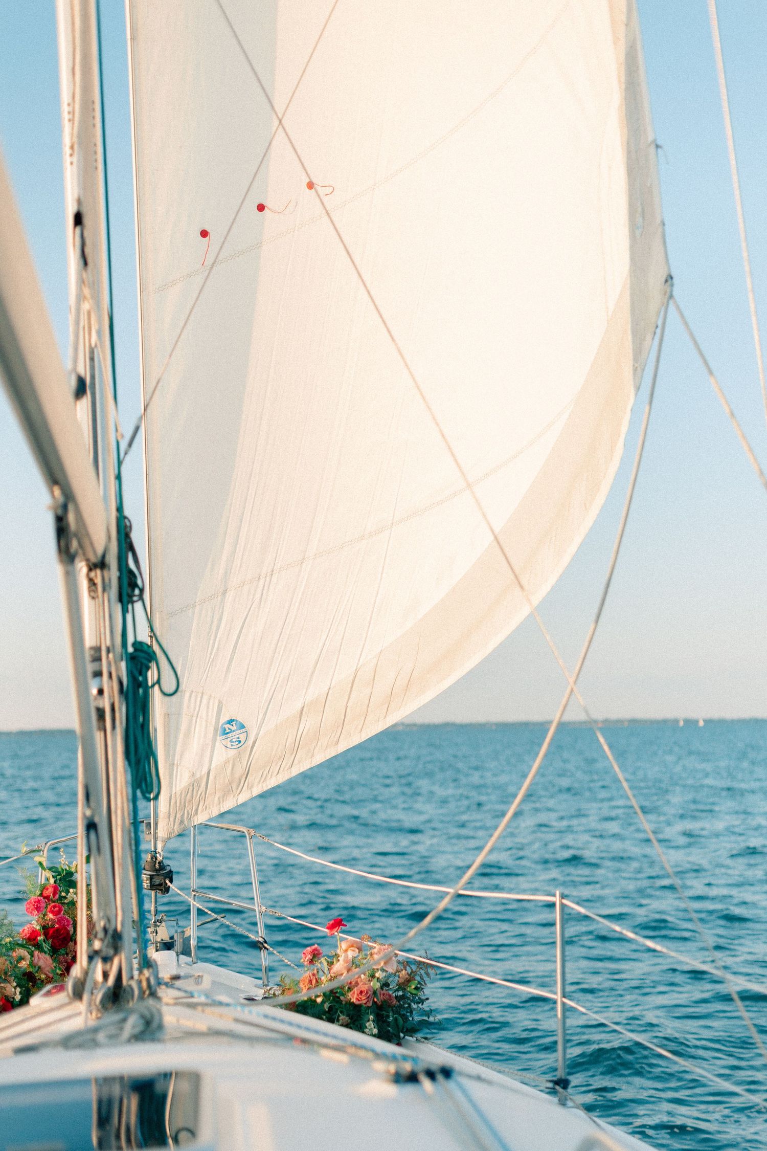 White sail billowing on a sailboat gliding across calm blue ocean waters with floral decorations on deck.