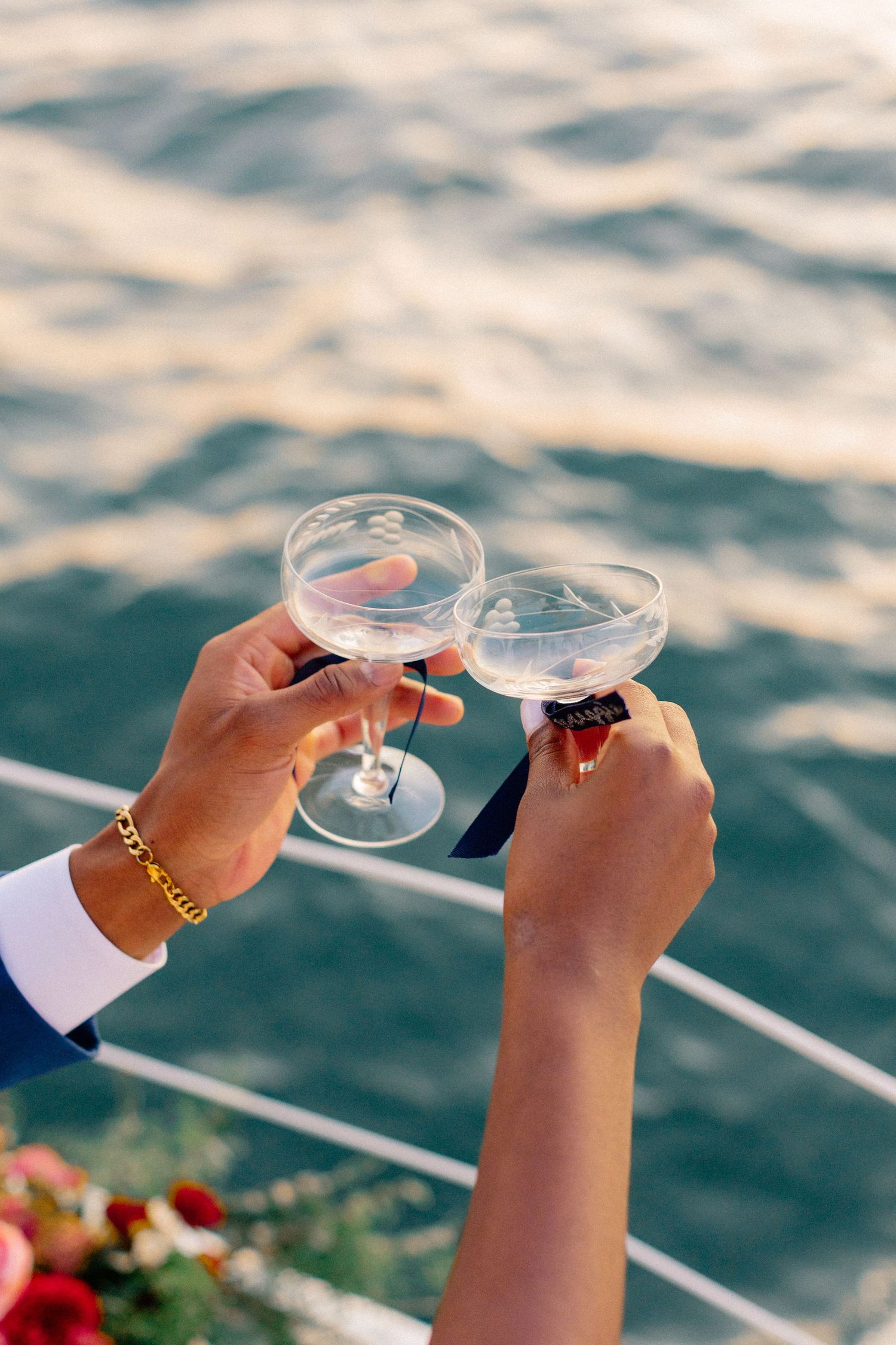 Couple toasting with coupe champagne glasses on a sailboat deck over sparkling ocean water.