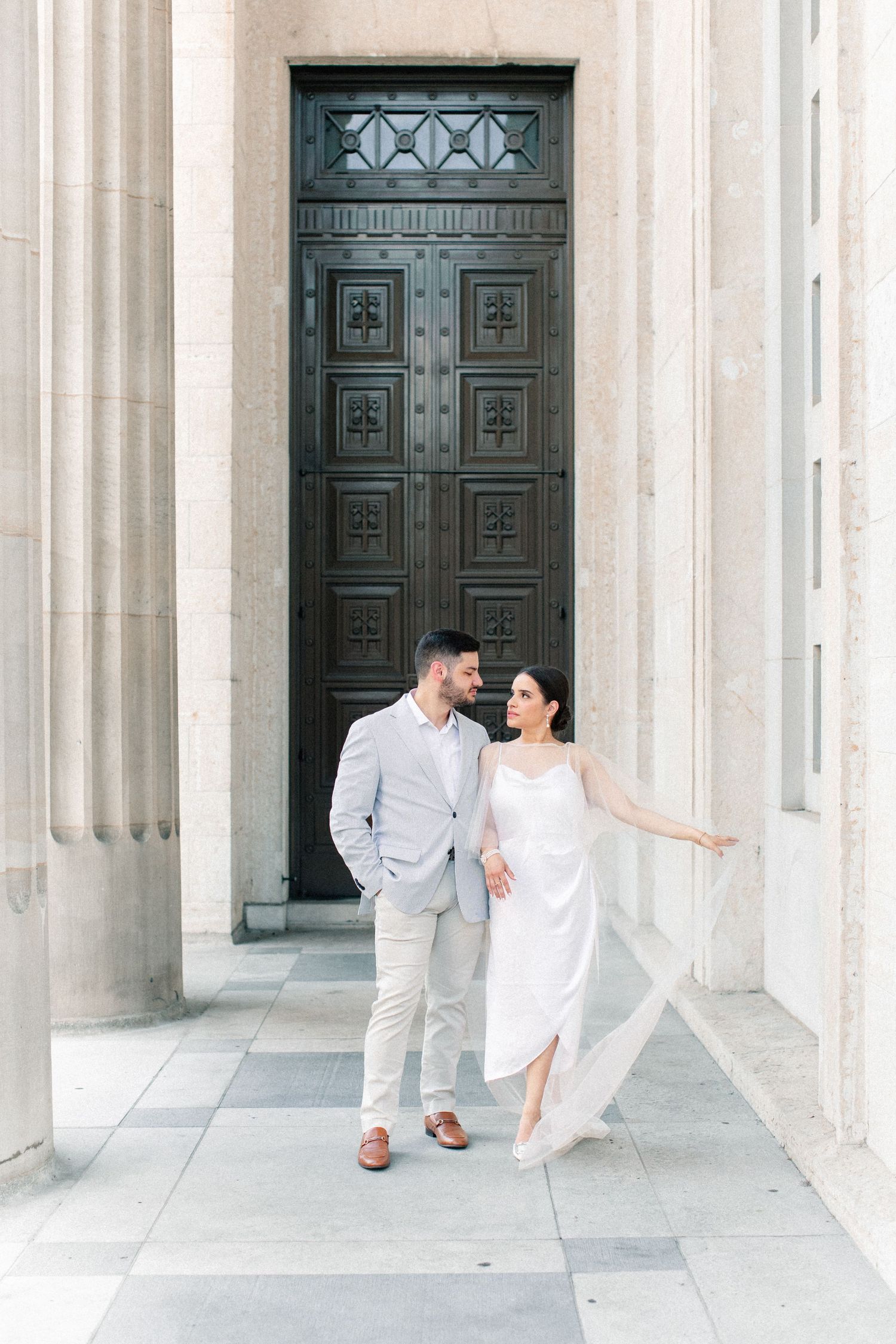 Couple in white dress and light suit walking hand in hand before ornate dark wooden doors flanked by stone columns.