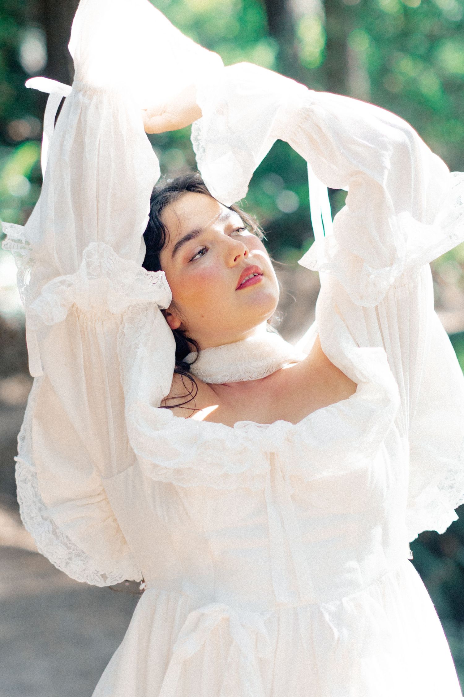 Woman in white ruffled long-sleeve dress lifts fabric overhead with eyes closed in sunlit garden setting.