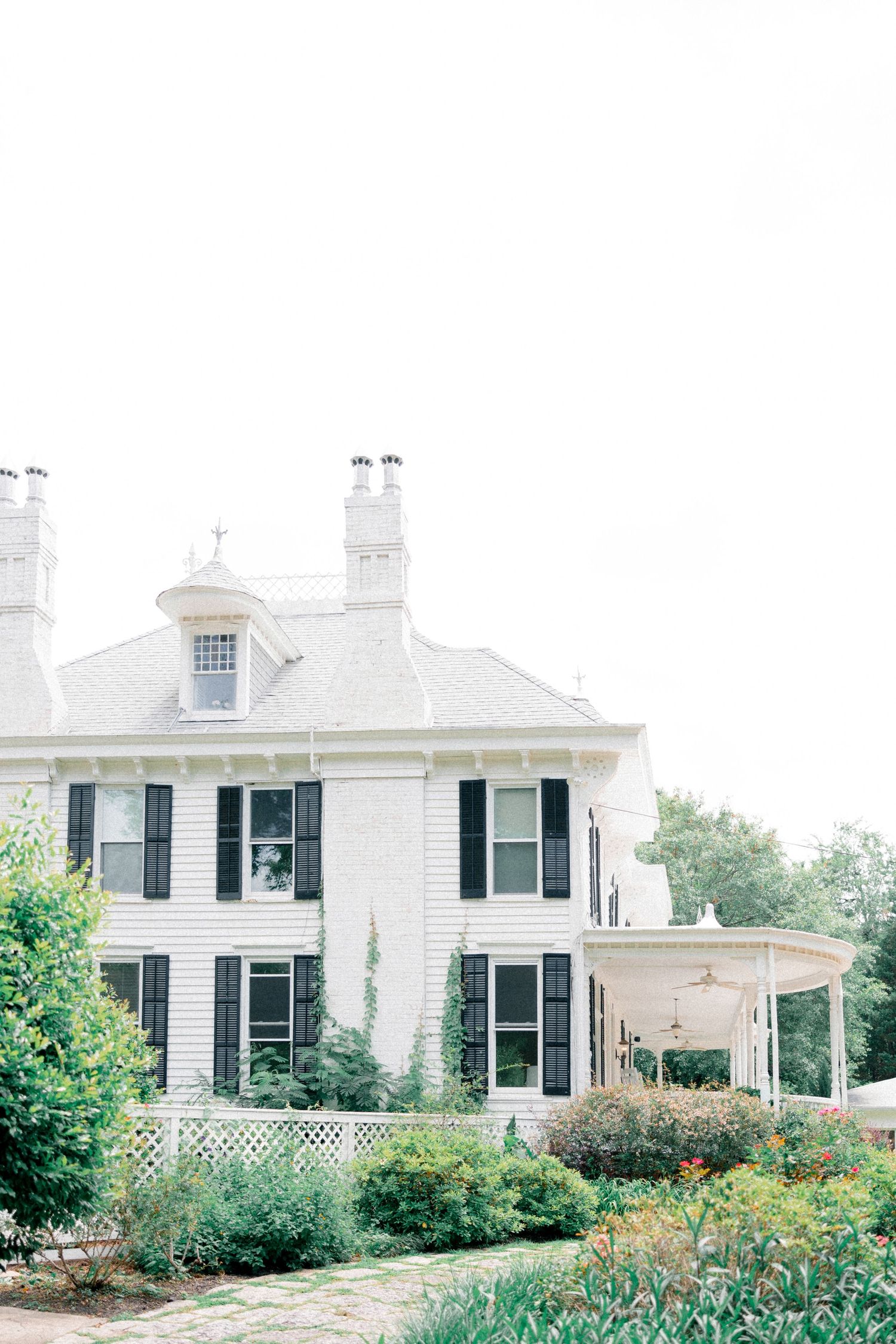Elegant white historic estate with black shutters and wraparound porch amid garden landscaping.