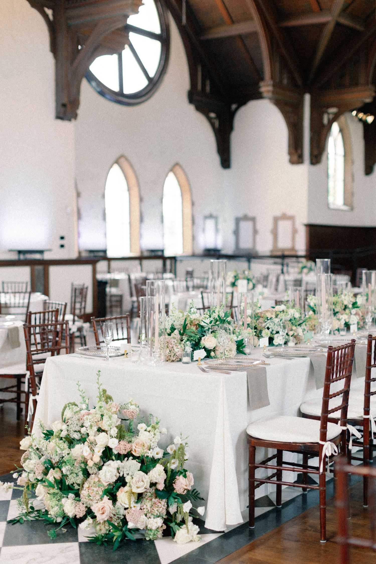 Grand wedding reception hall with Gothic arched windows, white floral centerpieces, and dark chiavari chairs.
