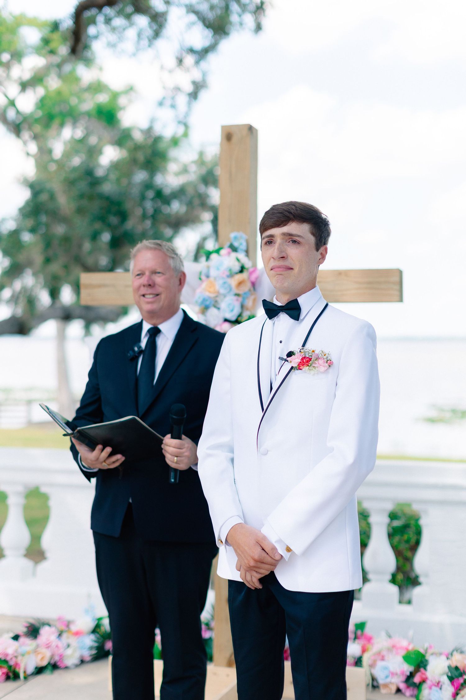 Groom in white tuxedo stands at outdoor wedding altar with officiant holding book and wooden cross backdrop, wedding at Bella Cosa