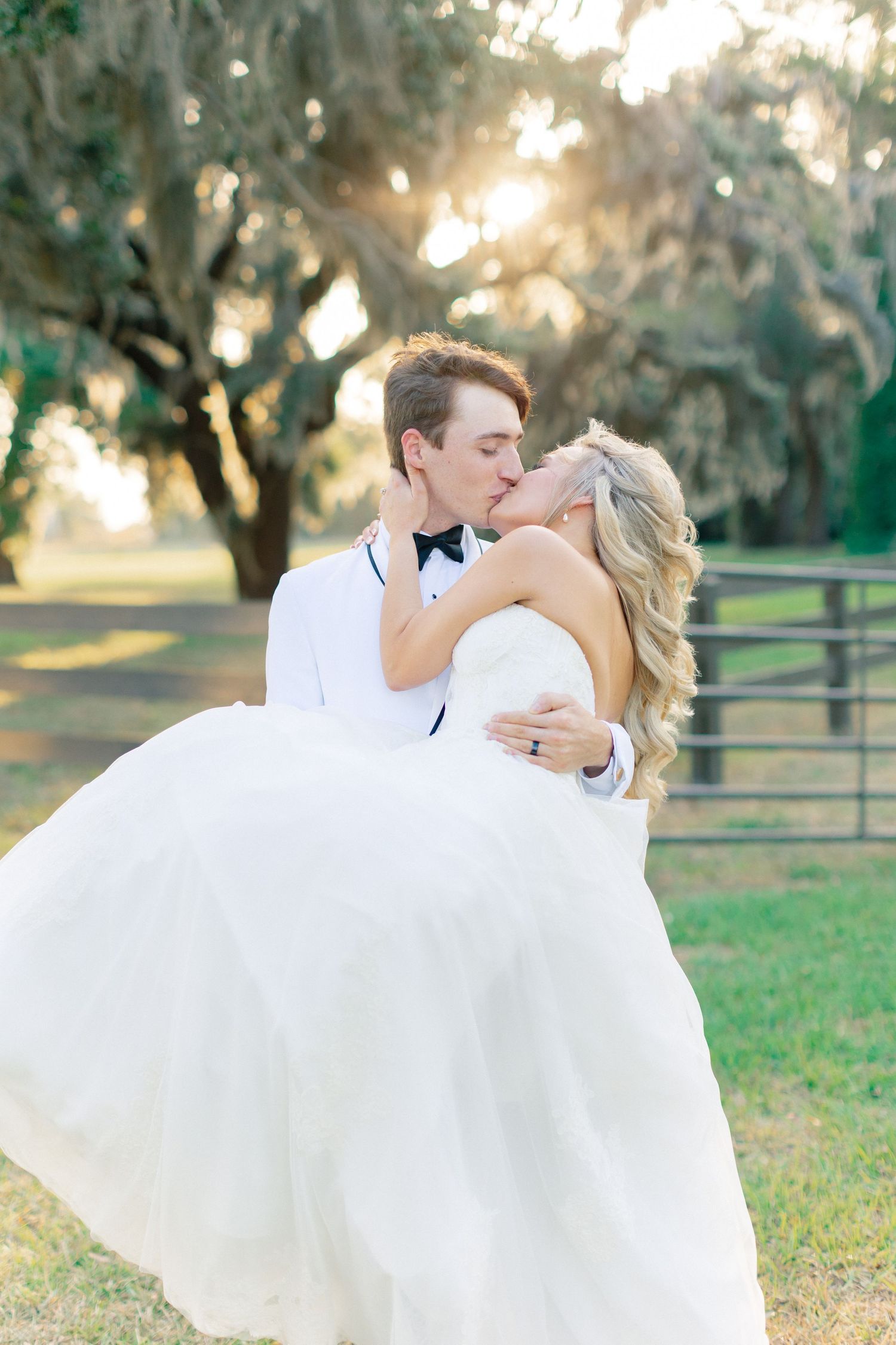 Groom in white tuxedo lifts and kisses bride in flowing white gown outdoors at golden hour near oak trees, wedding at Bella Cosa