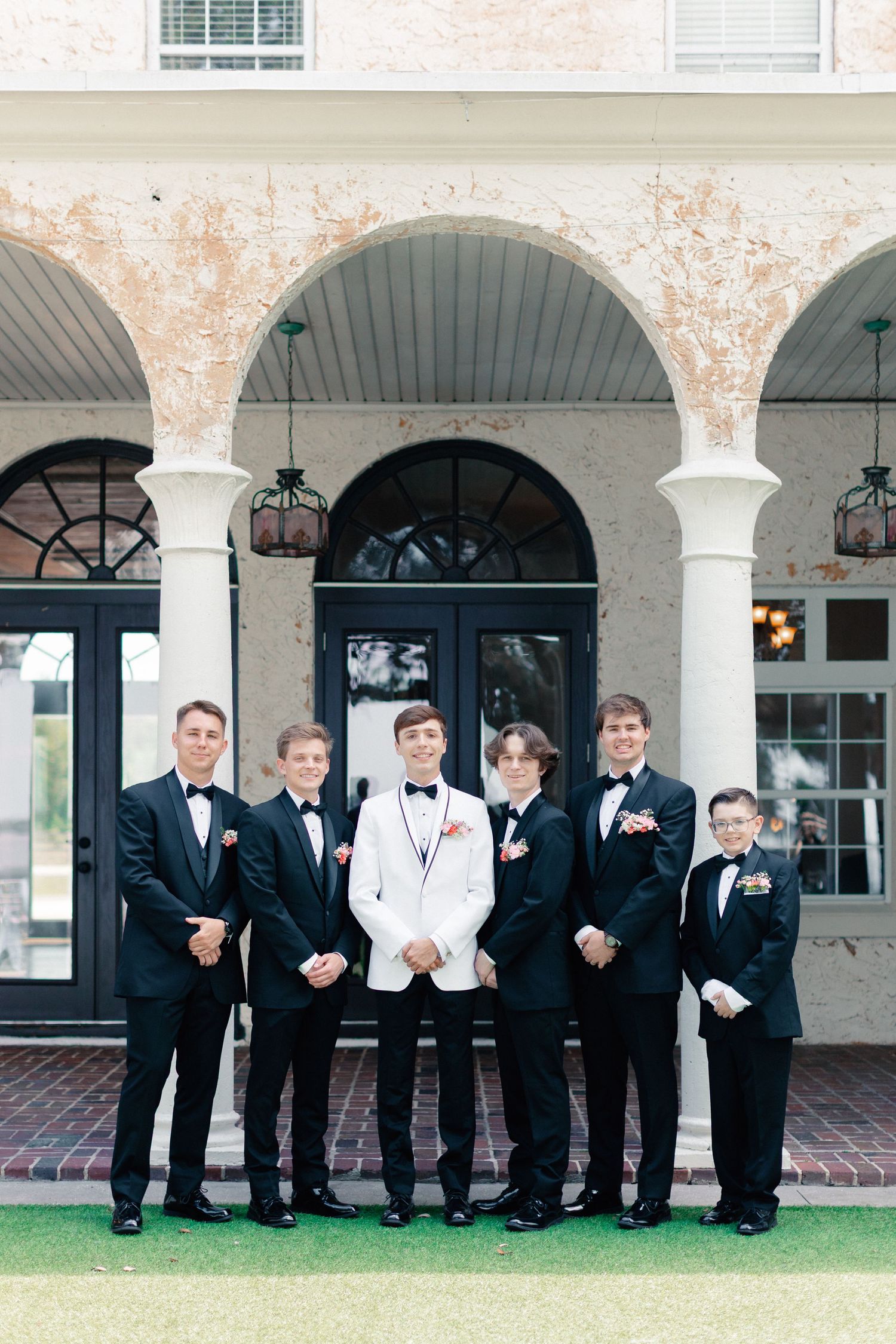 A groom in a white tuxedo stands with five groomsmen in black tuxedos outside an elegant venue with arched columns, wedding at Bella Cosa