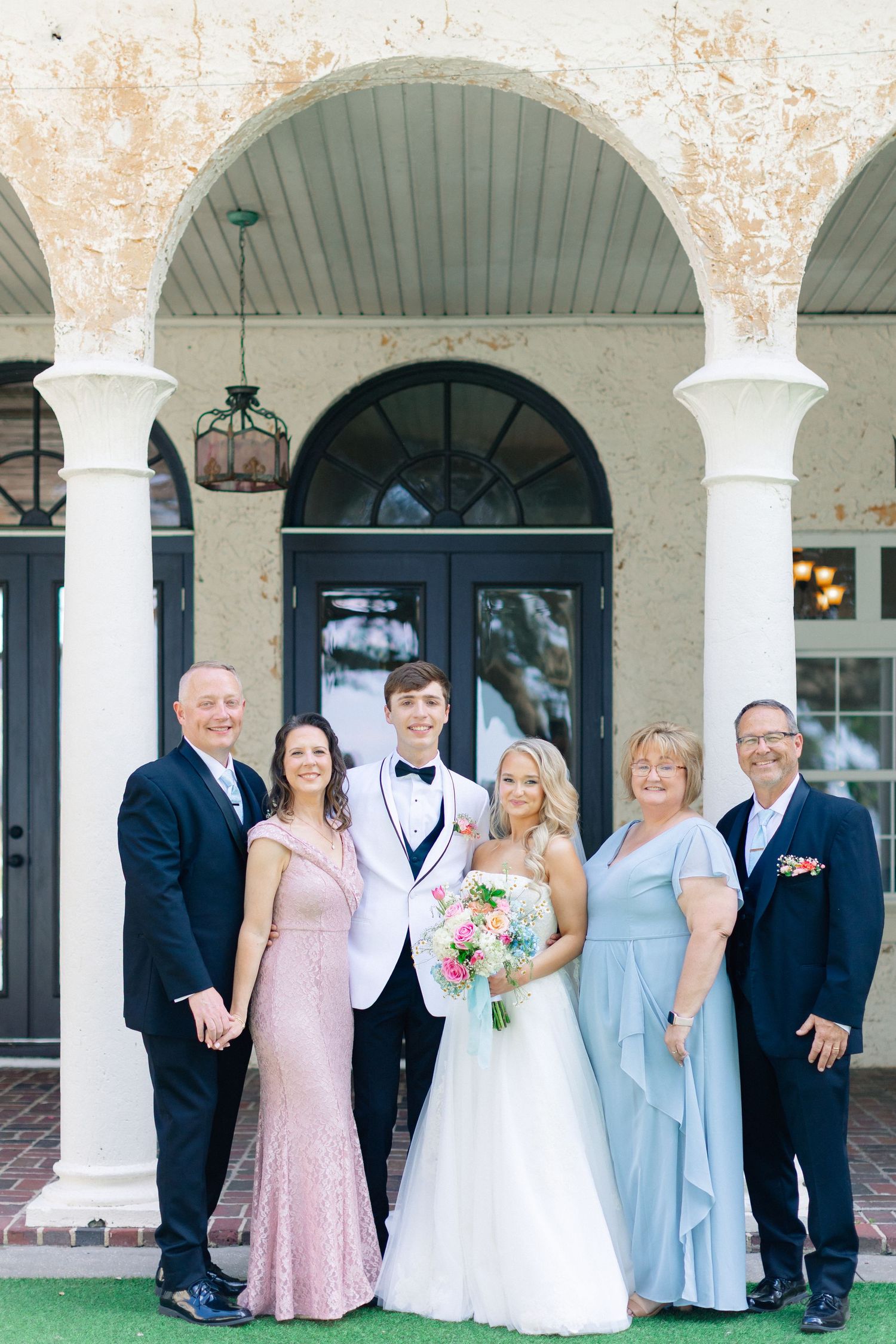 Wedding couple poses with family in formal attire before arched doorway of elegant venue, wedding at Bella Cosa