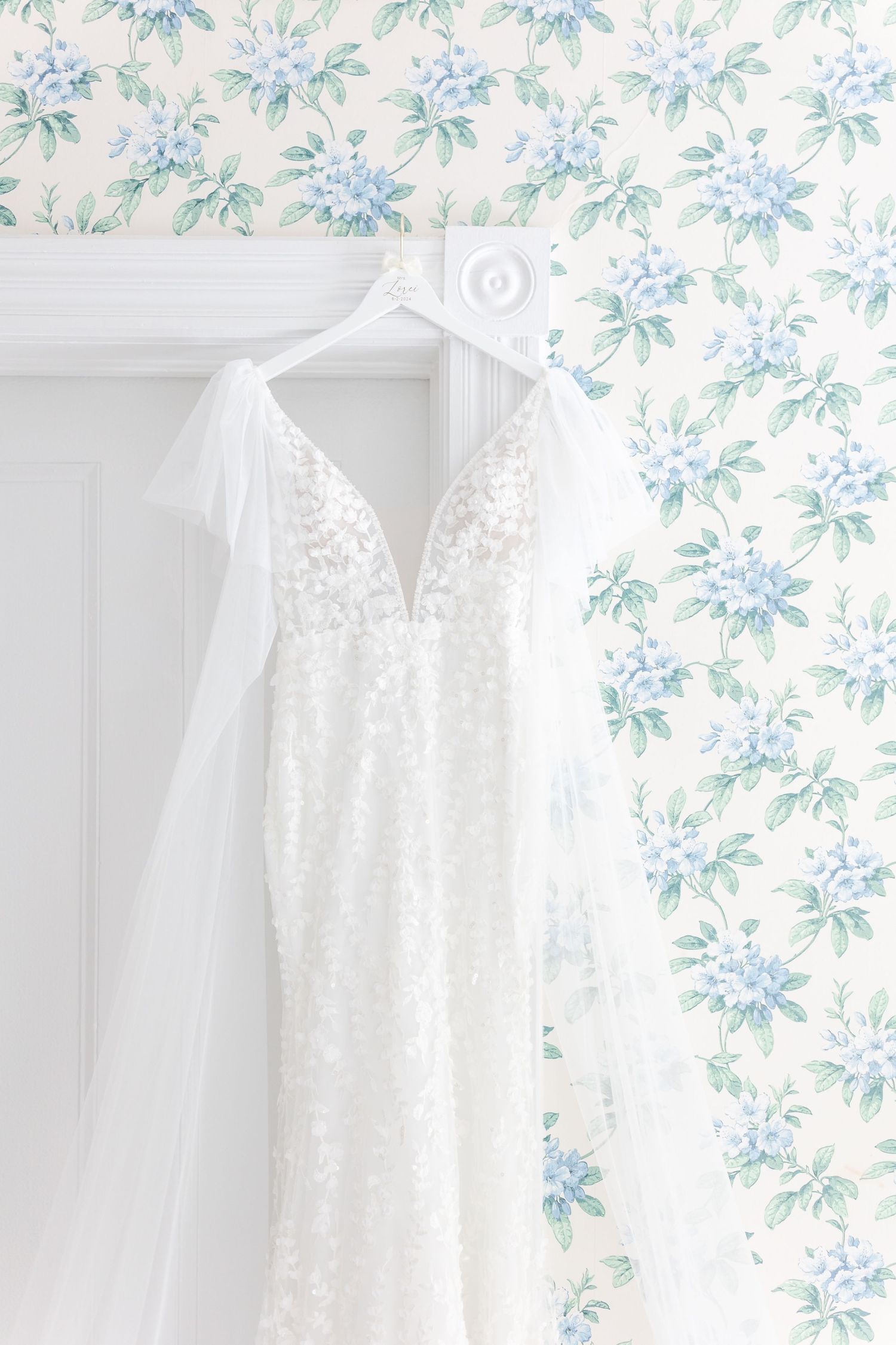 Elegant white lace wedding gown with deep V-neckline and flutter sleeves hanging on a hanger against floral wallpaper.