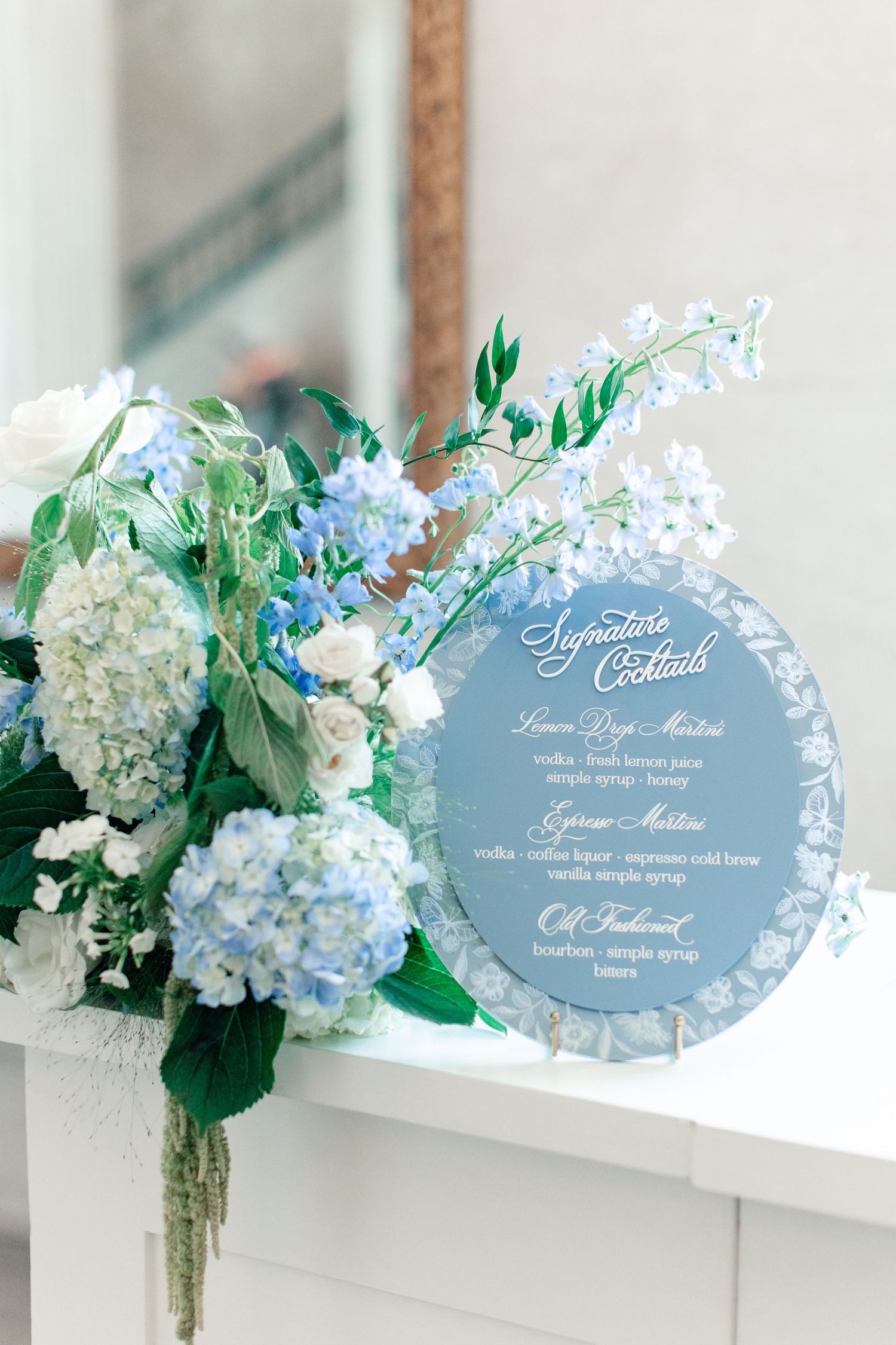 Blue acrylic signature cocktails sign beside elegant hydrangea and delphinium floral arrangement at wedding reception.
