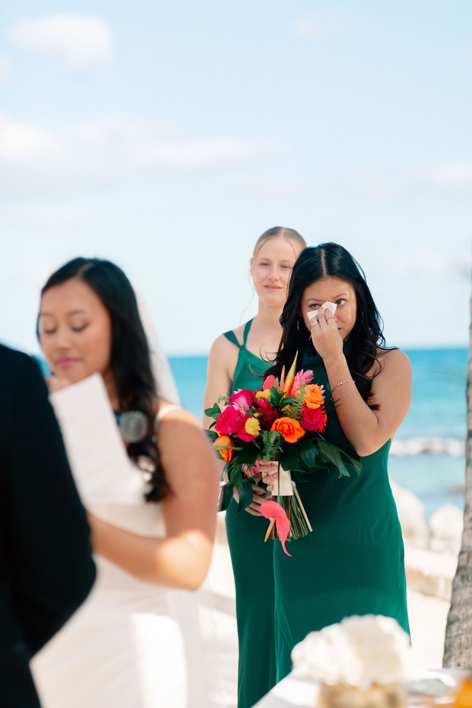 Emotional bridesmaid in green dress crying while holding colorful bouquet at outdoor beach wedding ceremony.