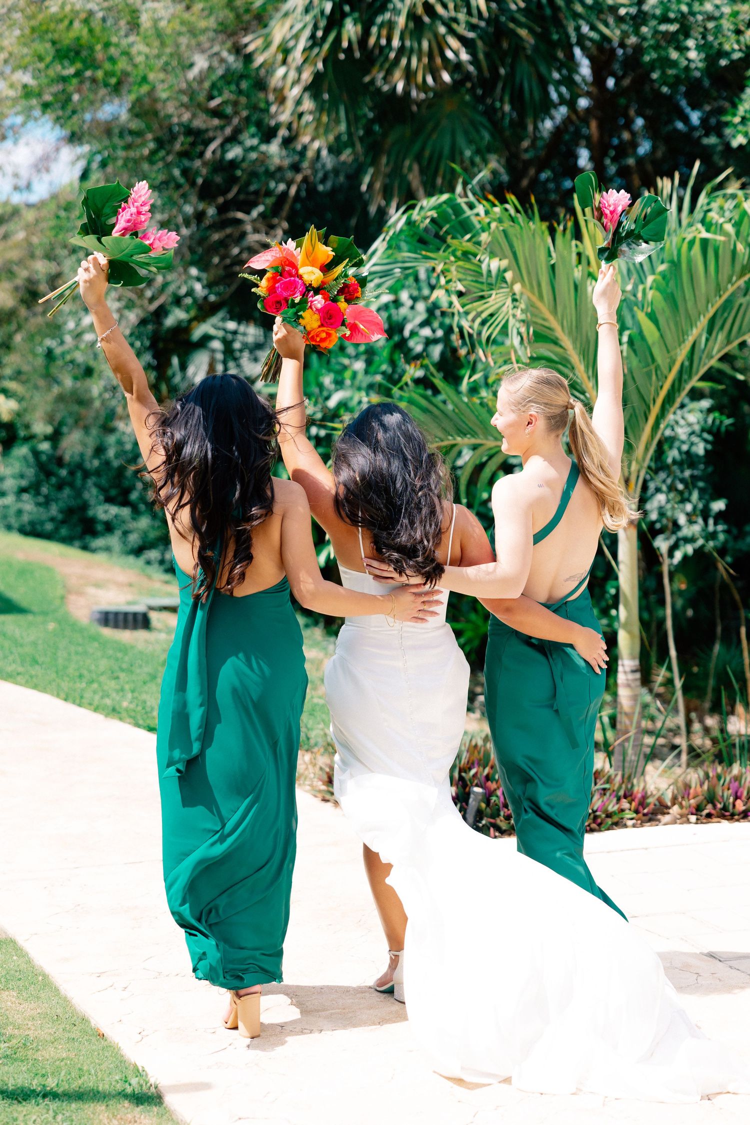 Bride and two bridesmaids in green gowns raising colorful bouquets while walking through tropical garden.