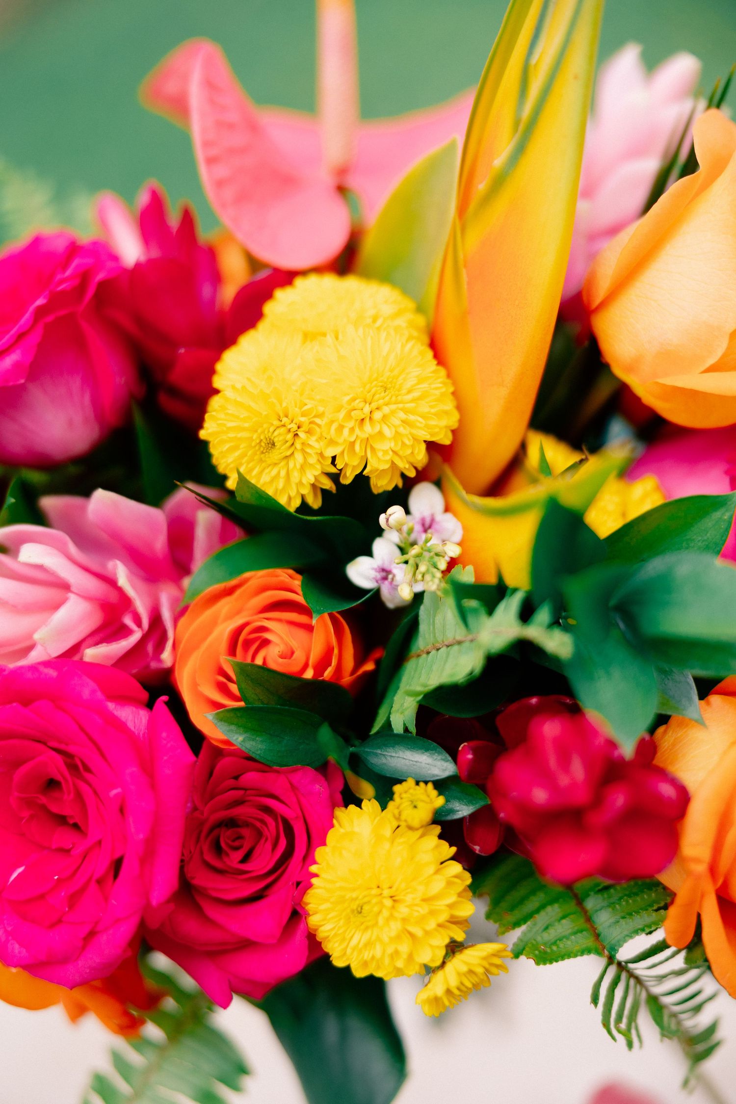 Vibrant tropical floral arrangement featuring hot pink roses, yellow chrysanthemums, orange blooms, and lush green foliage.
