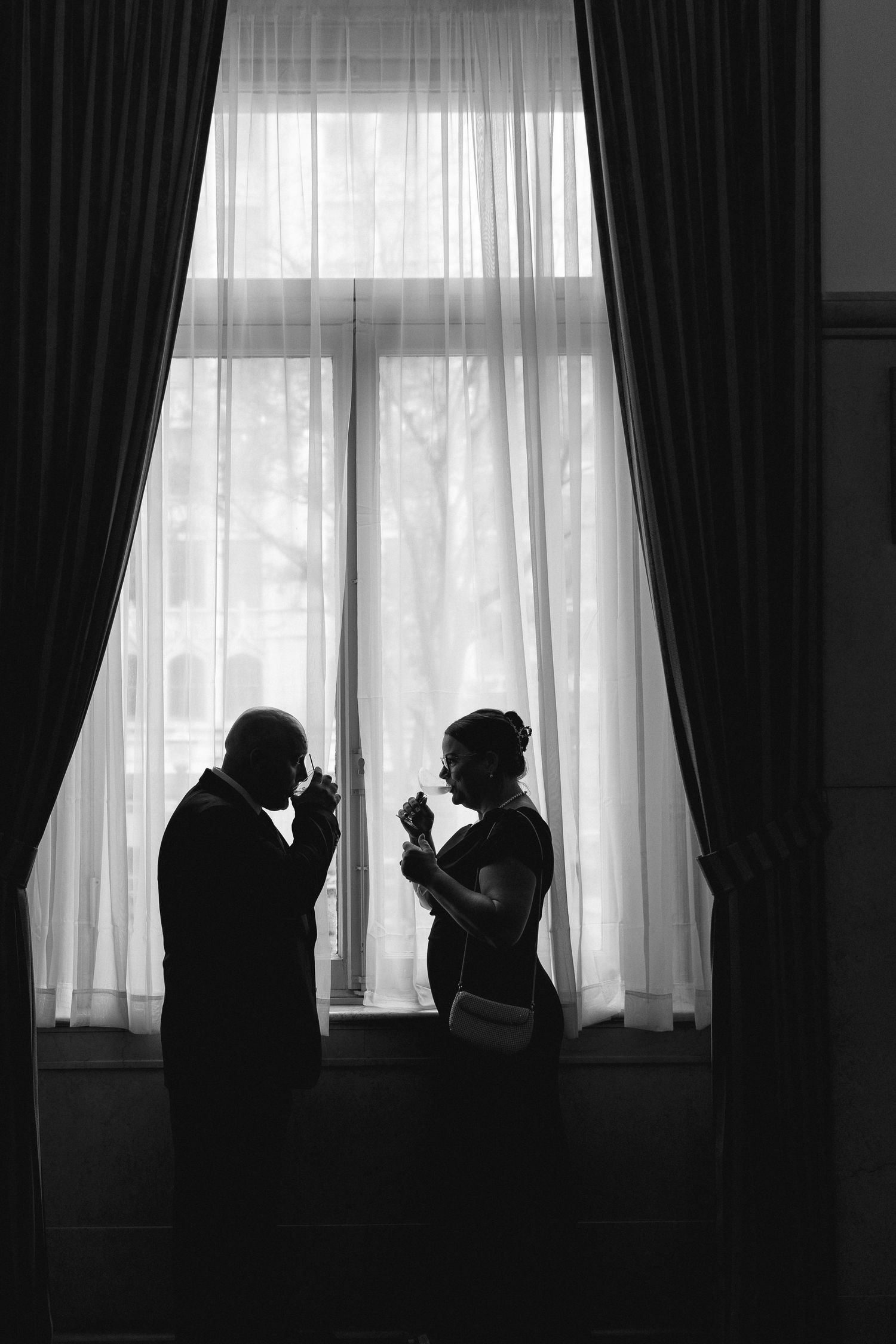 Black and white silhouette of elegant couple toasting with glasses by a large draped window.