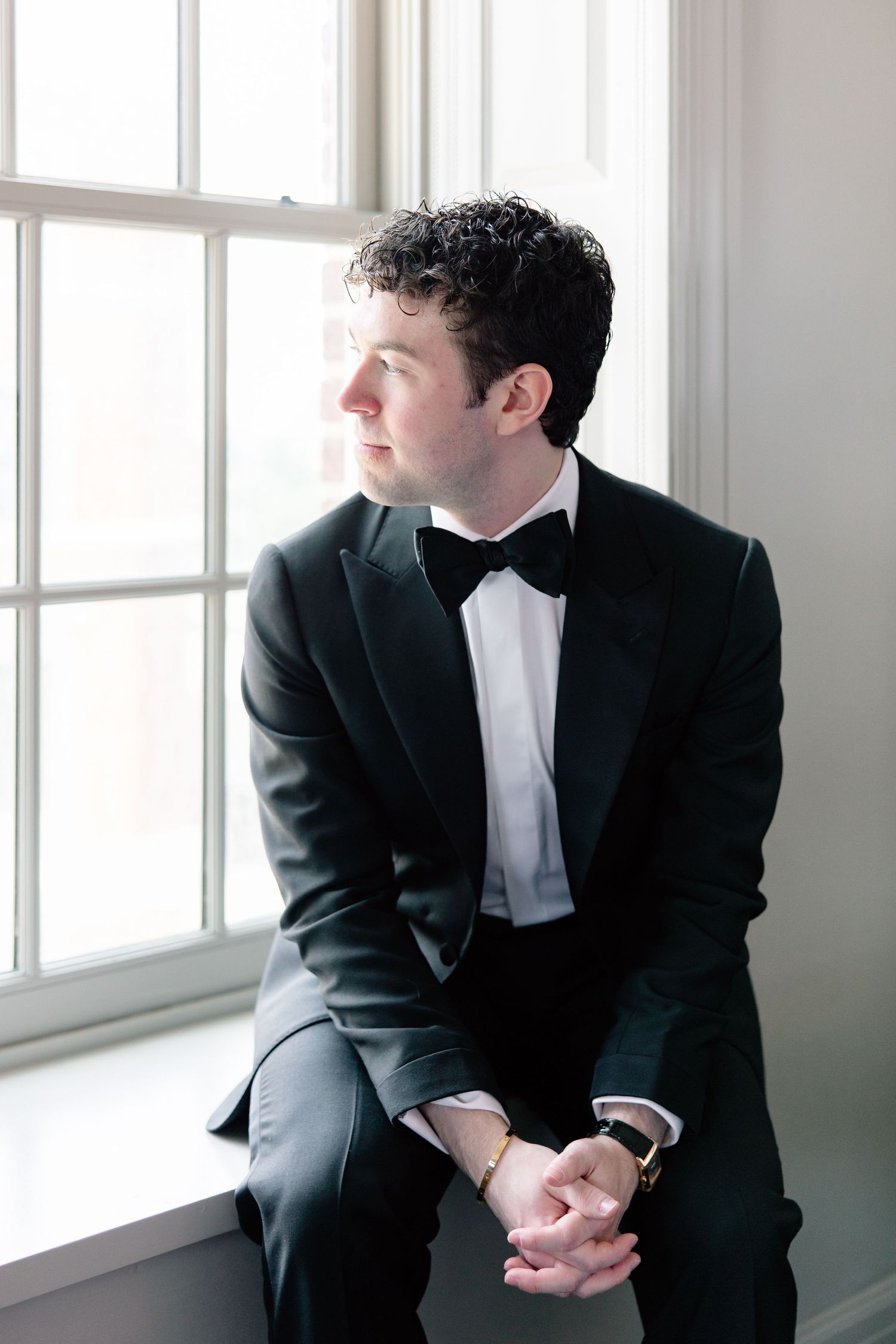 A dapper groom in a black tuxedo and bow tie sits by a window, gazing thoughtfully into the distance.
