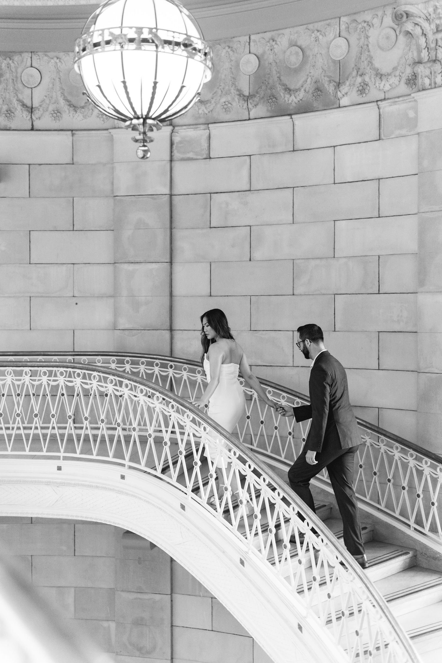 Black and white photo of bride and groom walking up a grand ornate staircase with decorative chandelier.