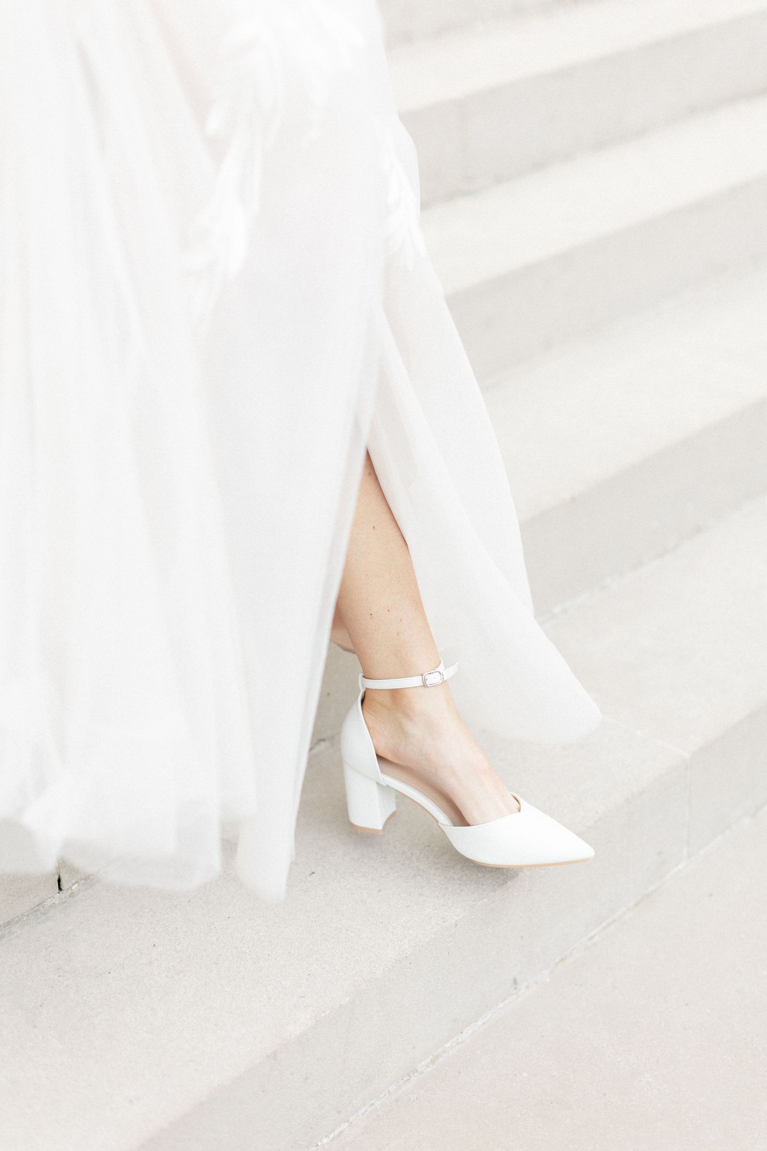 Close-up of a bride wearing elegant white block-heel ankle-strap shoes on white marble steps.