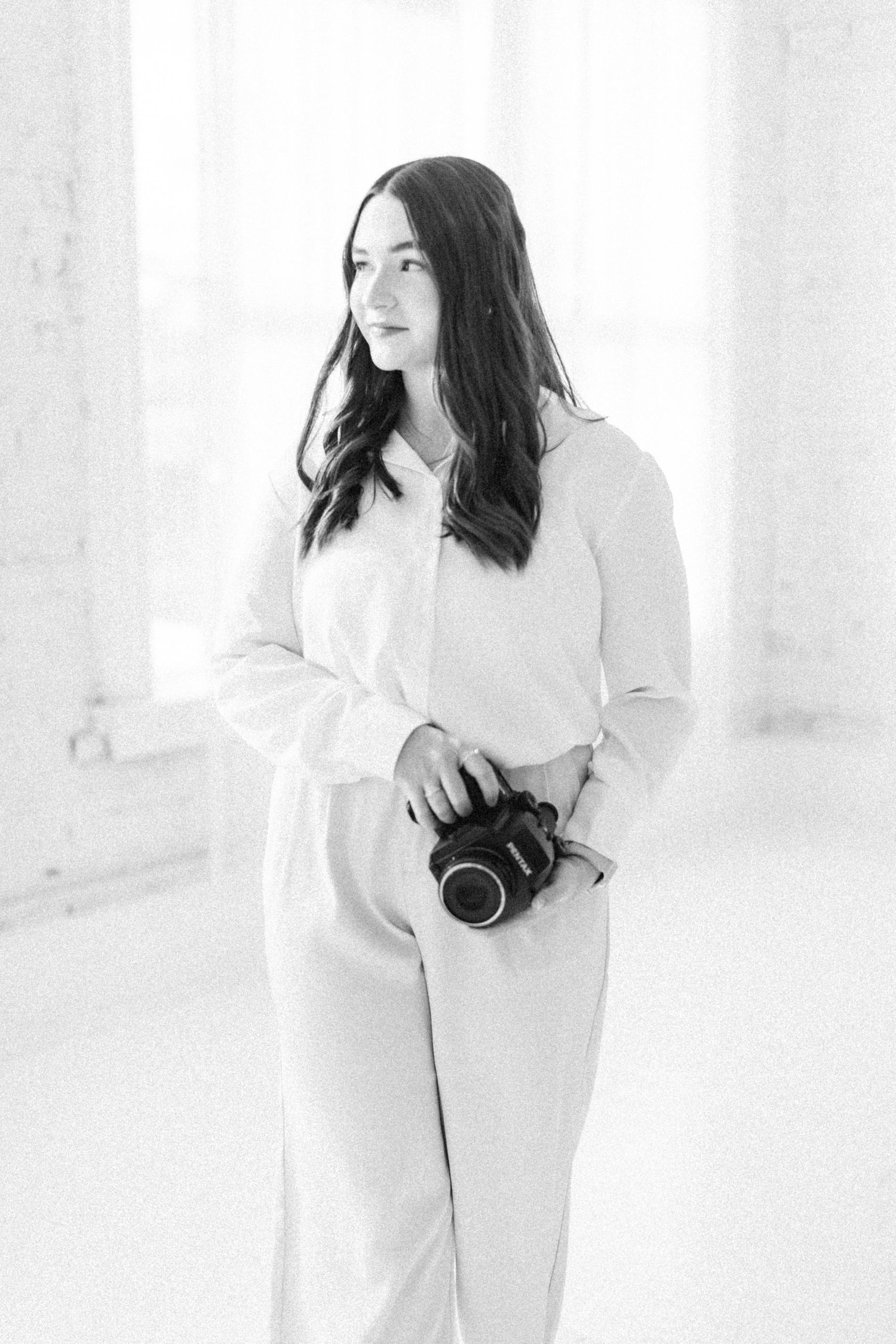 Black and white portrait of a female photographer holding a camera in a bright white studio.