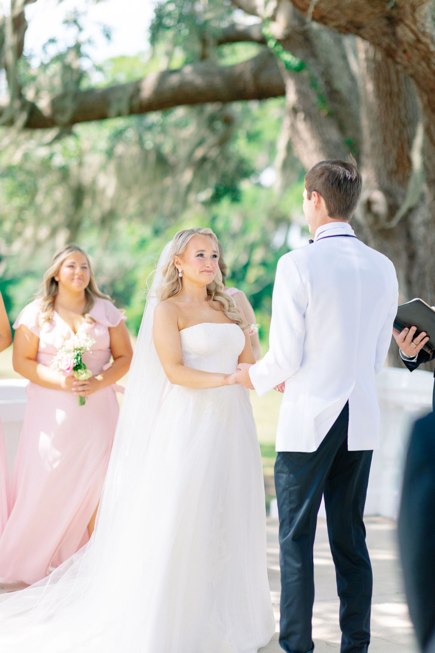 Bride in strapless gown exchanges vows with groom in white jacket at outdoor wedding ceremony with bridesmaids in pink, wedding at Bella Cos