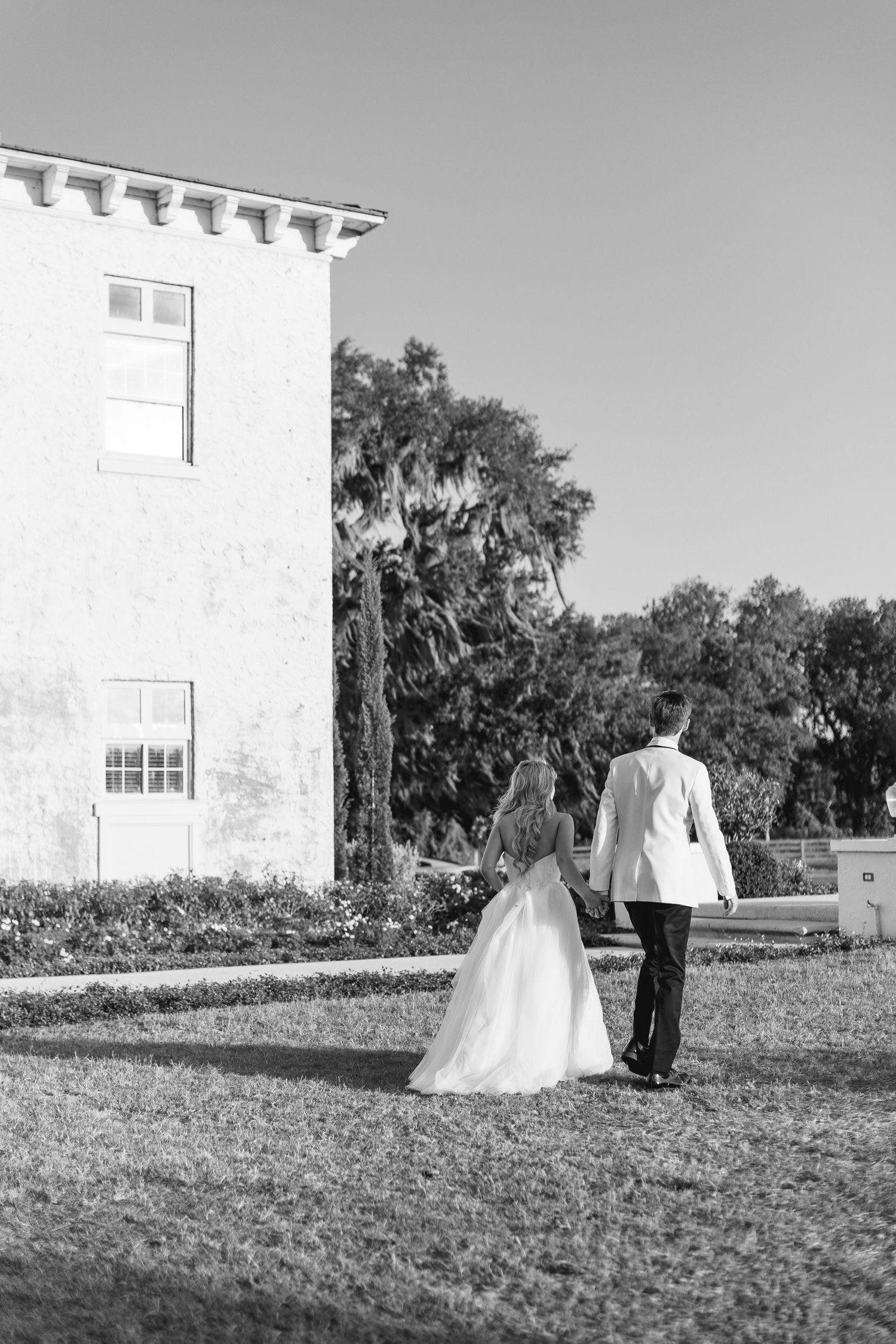 Black and white photo of a bride and groom walking hand in hand toward an elegant white building surrounded by lush greenery, Florida