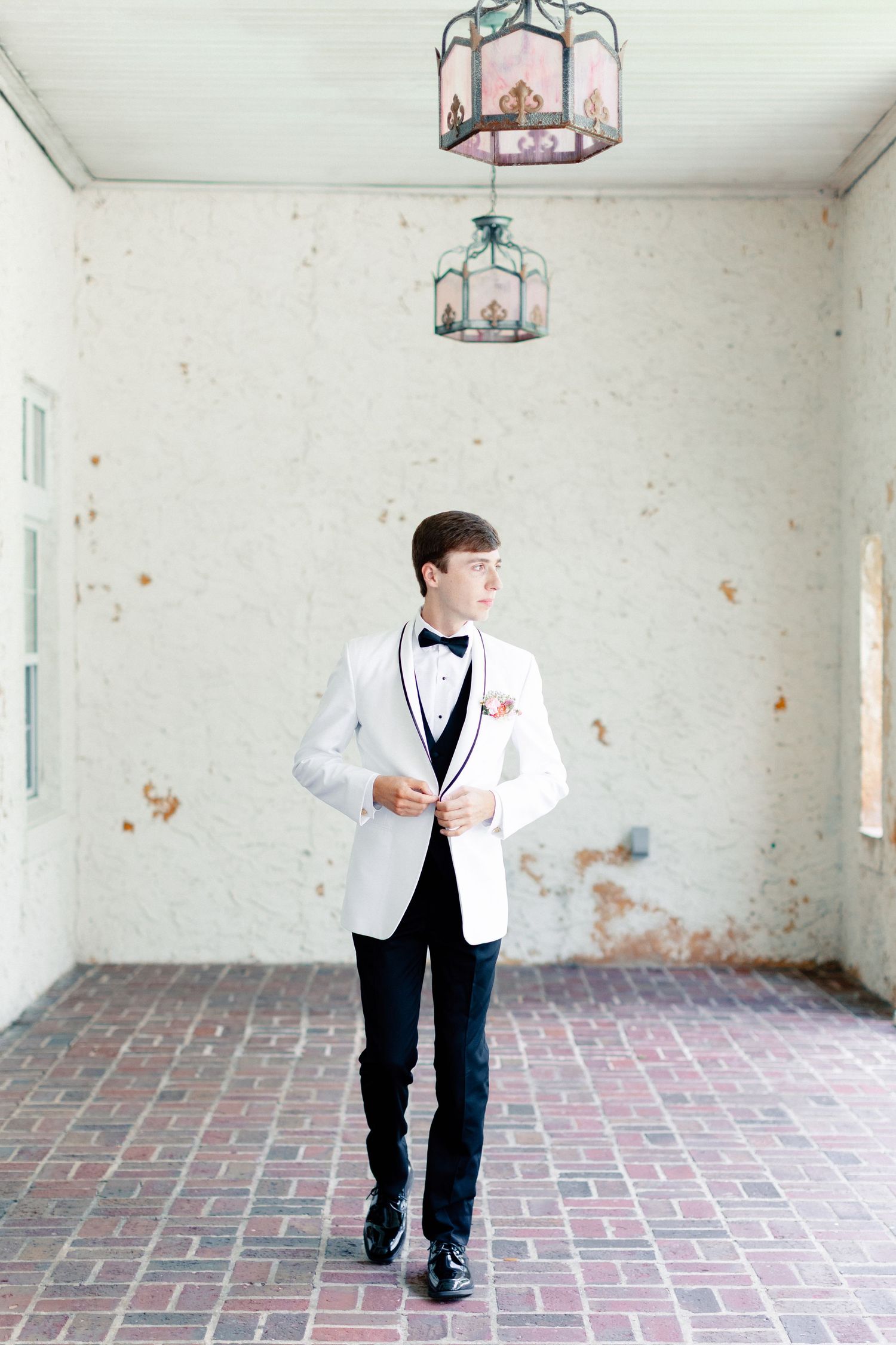 Young groom in white tuxedo jacket and black bow tie buttoning his jacket in an elegant venue, wedding at Bella Cosa