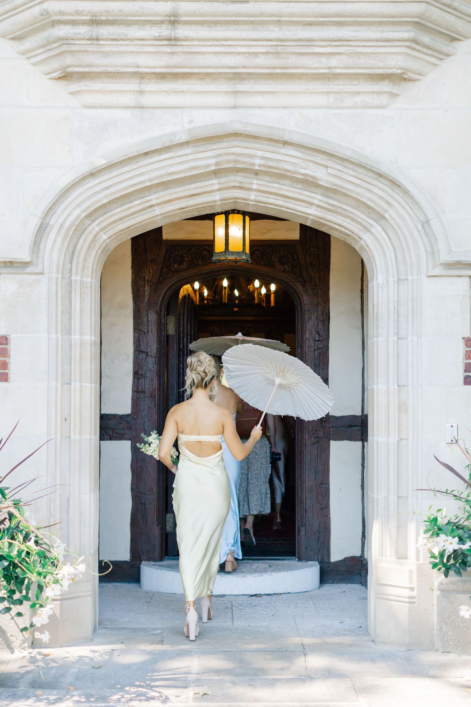 Bridesmaid in sage slip dress carries white parasol through a grand stone arched entrance.