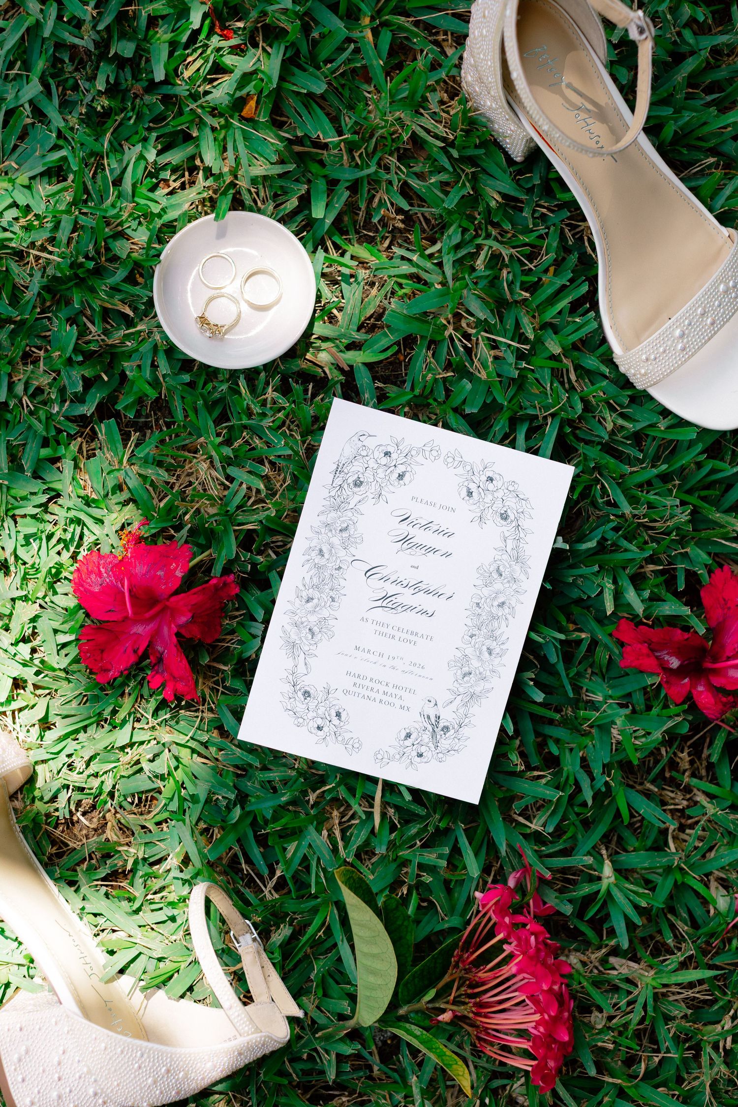 Wedding flat lay with floral invitation, gold rings, red hibiscus flowers, and white heeled bridal shoes on green grass.