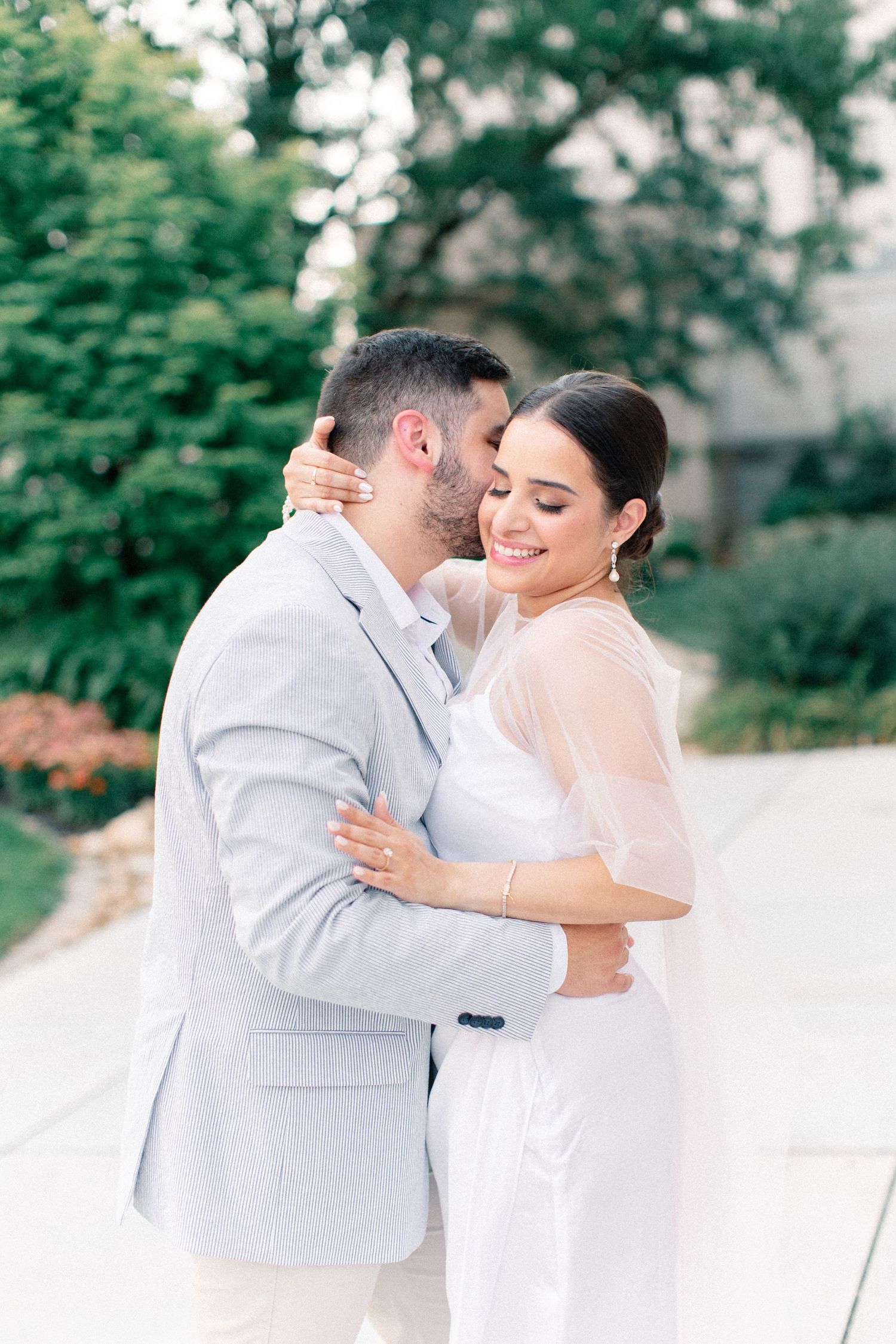 Couple embracing outdoors, groom in light gray suit kissing bride's cheek as she smiles, wearing white outfit.