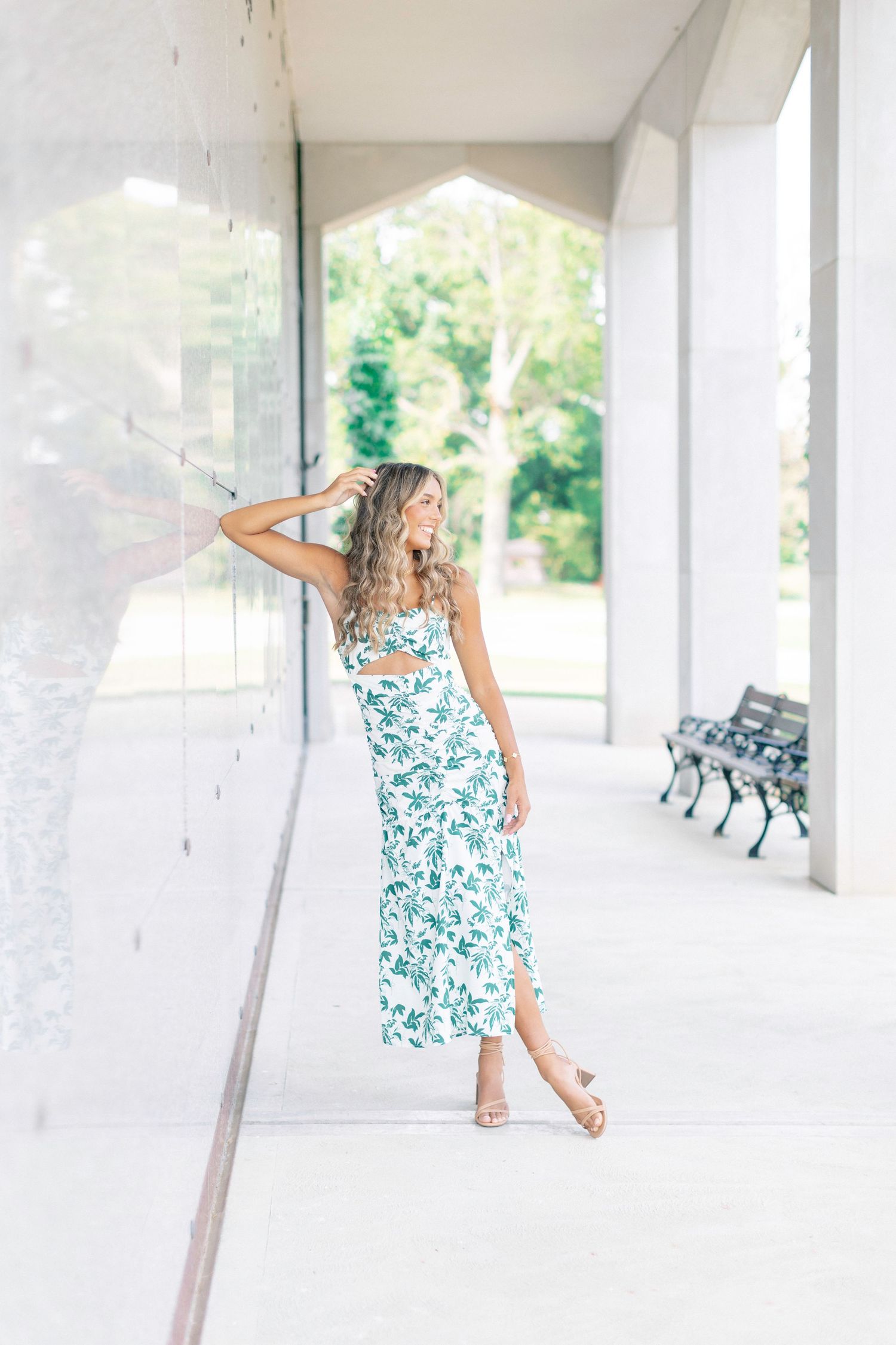 Woman in floral white and green midi dress posing outdoors near glass wall, wearing gold heeled sandals.