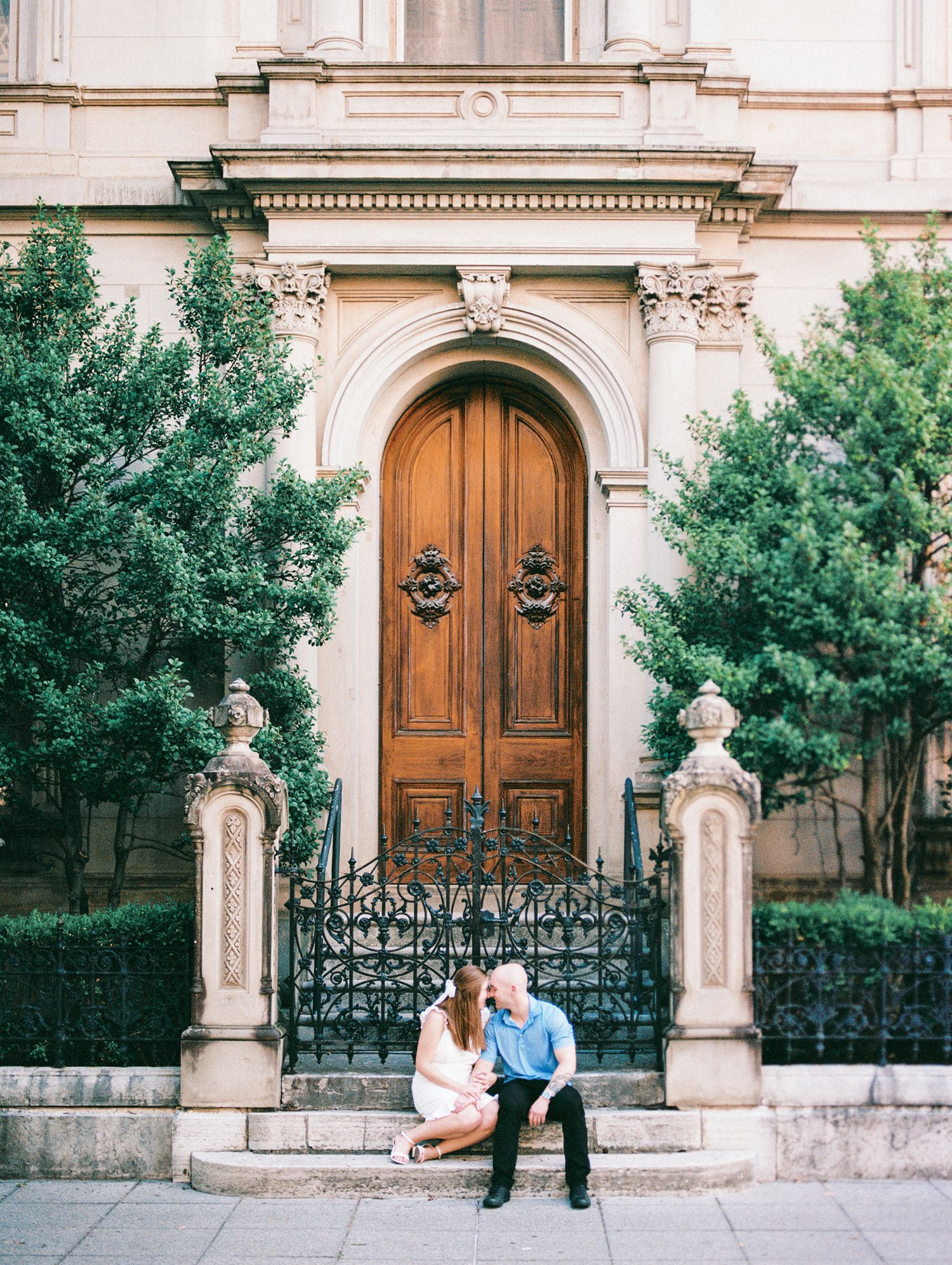 Couple sitting on steps before ornate wooden arched door of historic building with iron gates and lush green trees.