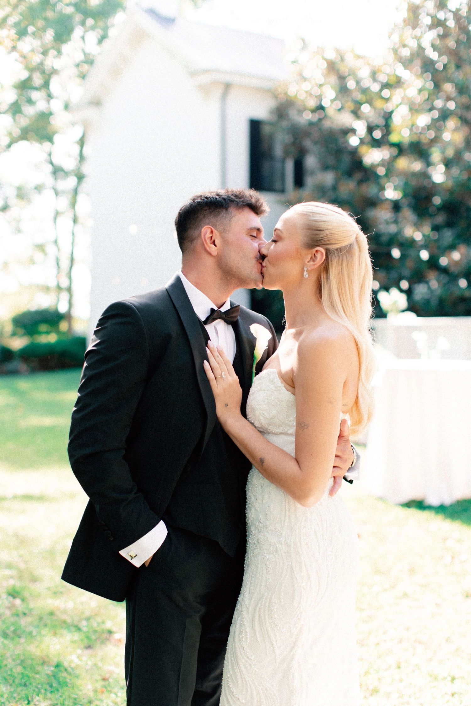Bride and groom share a romantic kiss outdoors, she in a beaded white gown, he in a classic black tuxedo.