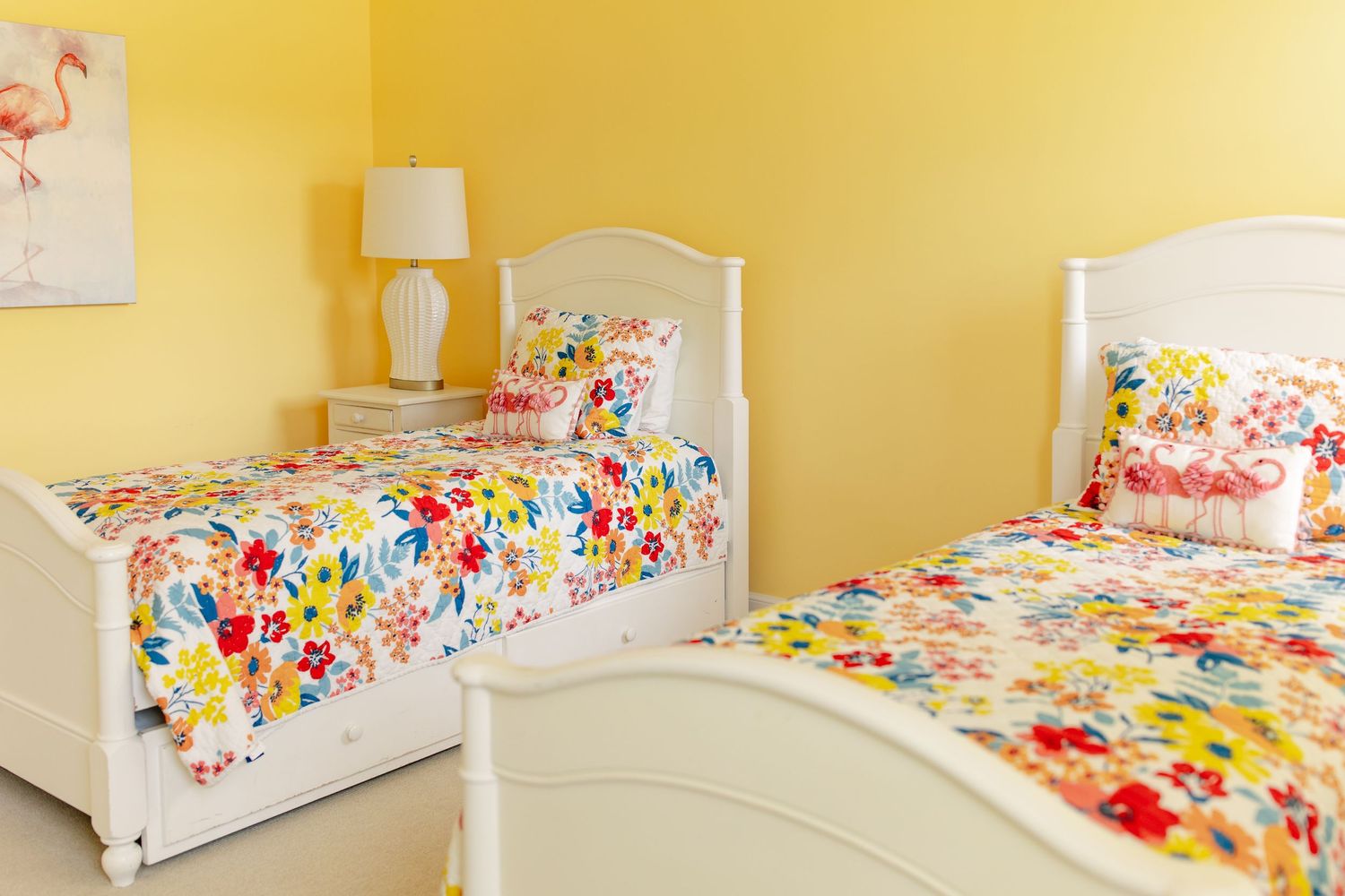 Bright yellow bedroom with two twin beds featuring colorful floral bedding and white furniture, accented by a flamingo wall art.