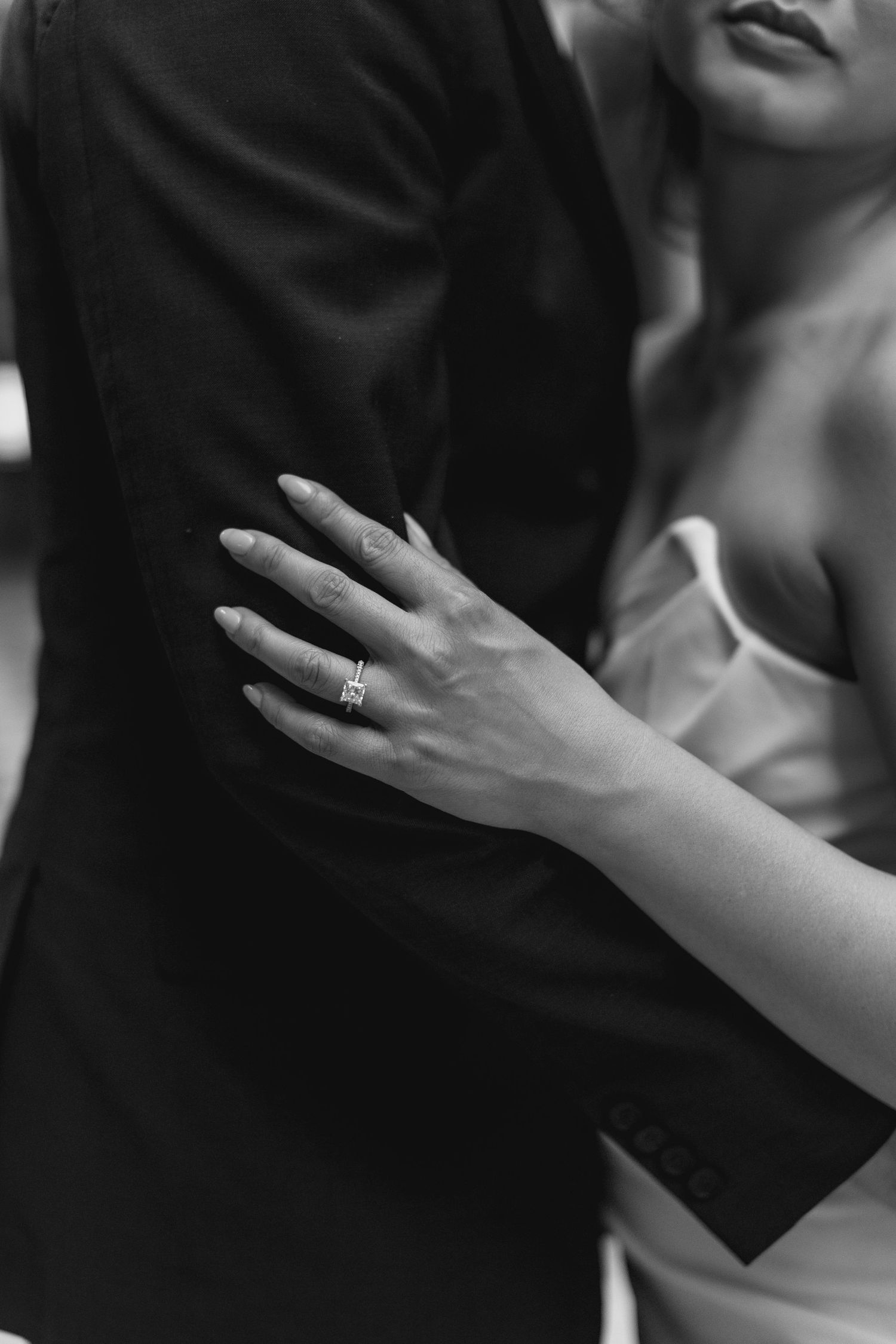 Black and white photo of a bride resting her hand on groom's suit, showcasing her diamond engagement ring.