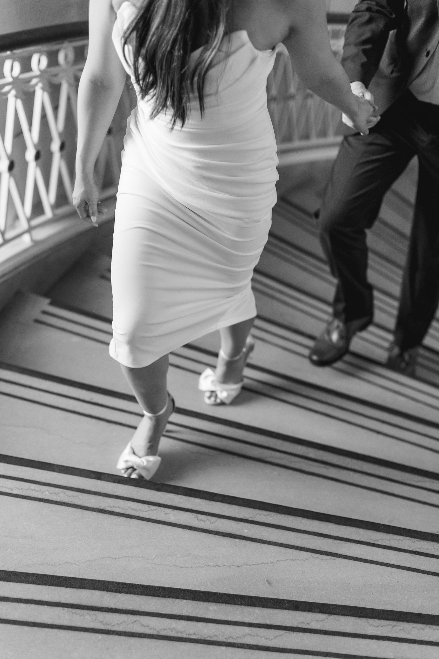 Black and white photo of bride in white ruched dress and heels walking up marble stairs with groom.