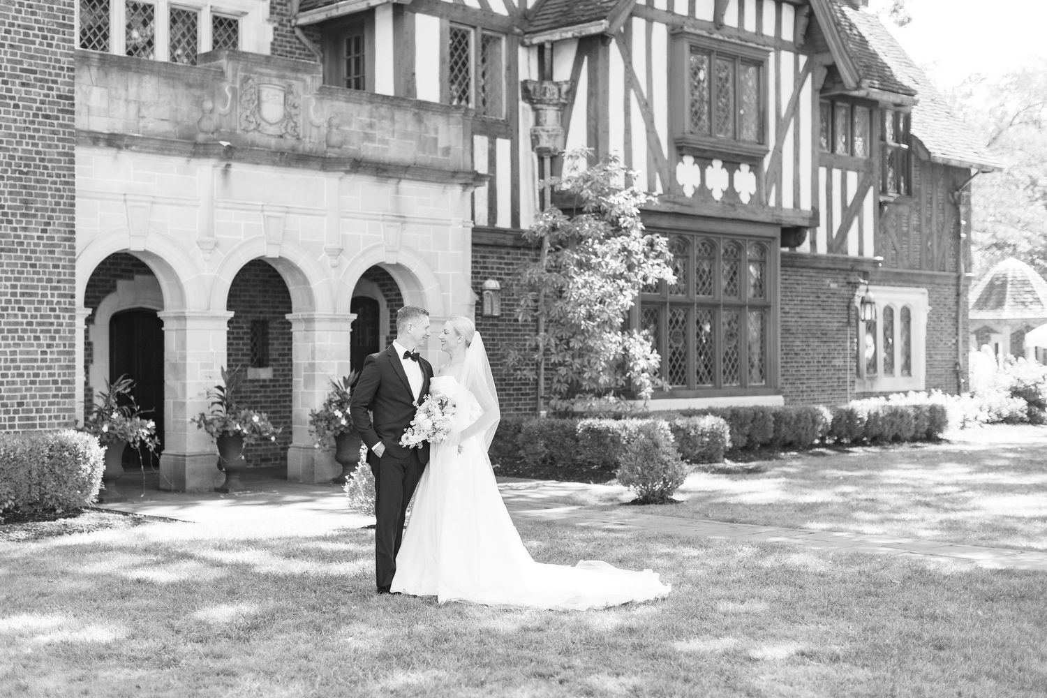 Black and white photo of bride and groom posing before a grand Tudor-style mansion on their wedding day.