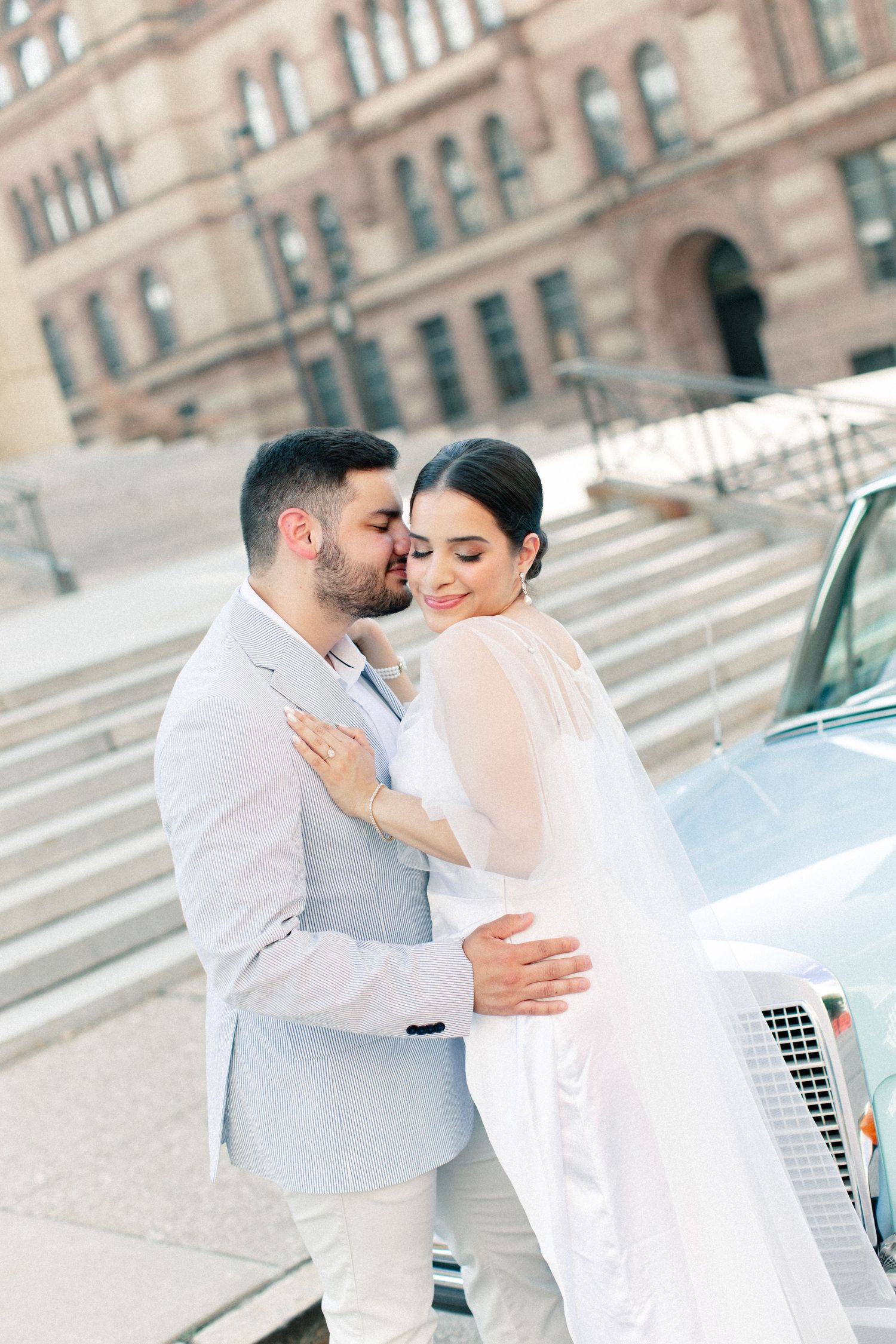 A groom in a light blue suit kisses his bride in a white gown near a classic car outside a historic building.