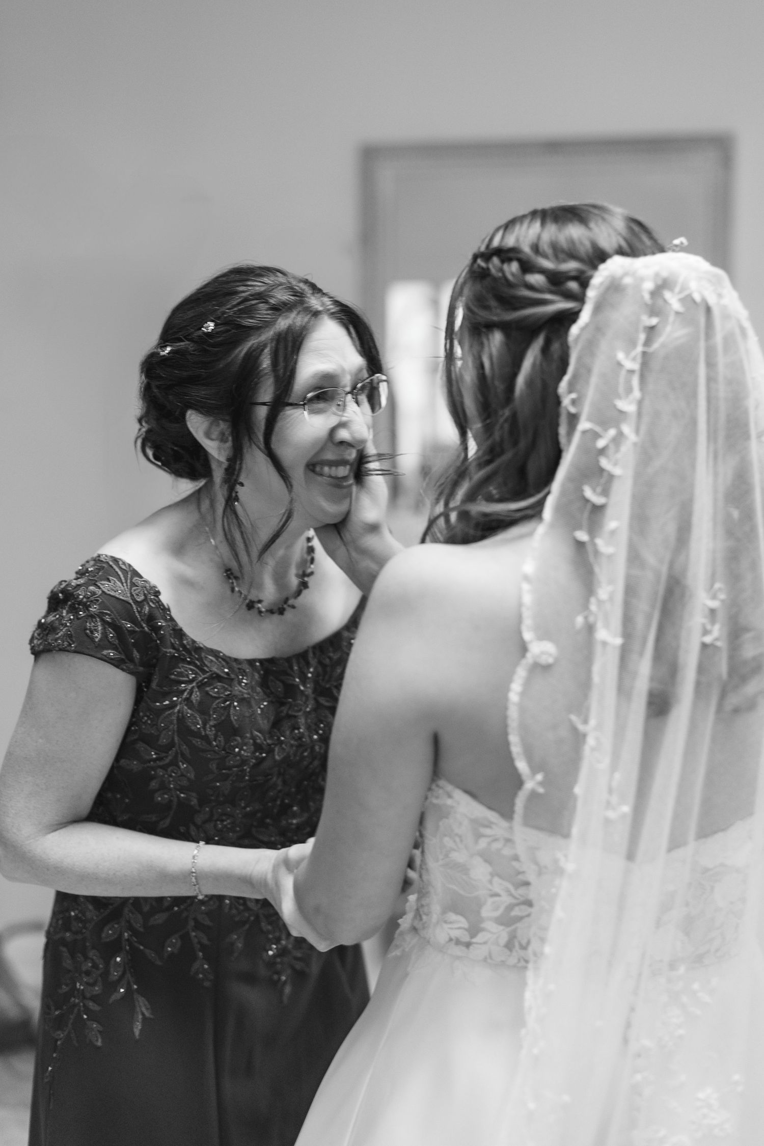 Black and white photo of emotional mother laughing with bride in lace veil before wedding ceremony.