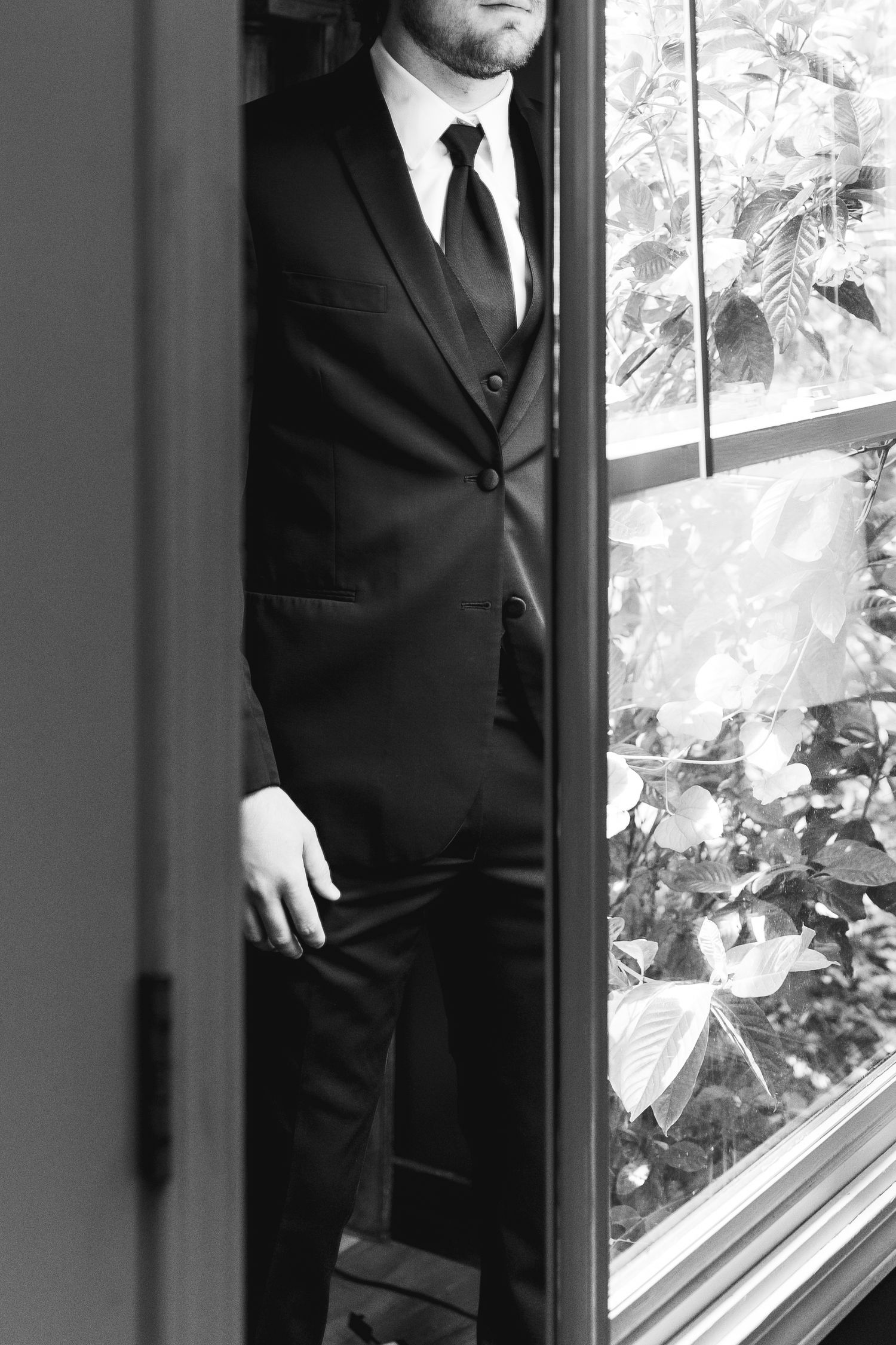 Black and white photo of a man in a black suit and tie standing by a window with foliage backdrop.