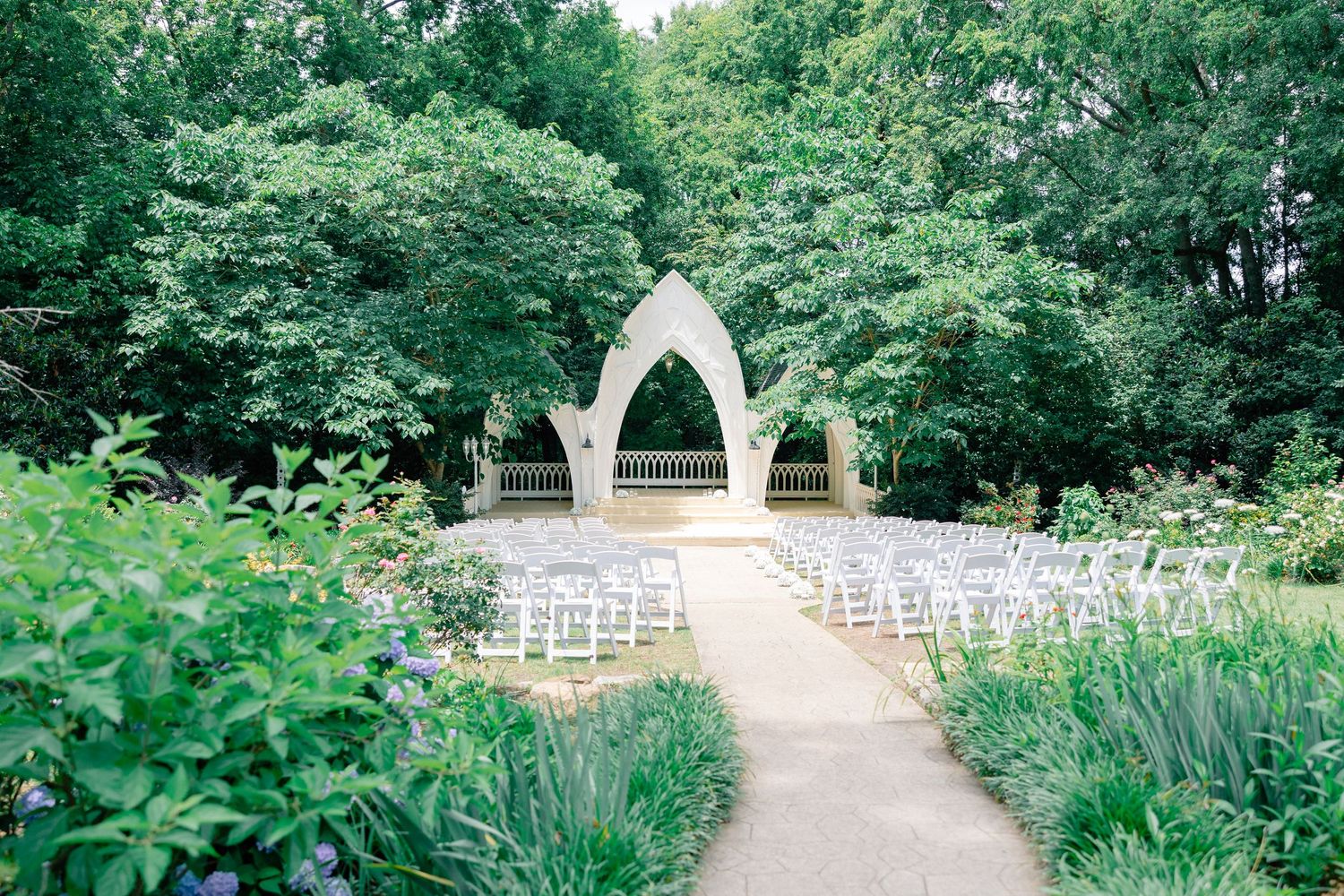 Outdoor wedding ceremony setup with white chairs along a garden path leading to an elegant white arch surrounded by lush green trees.